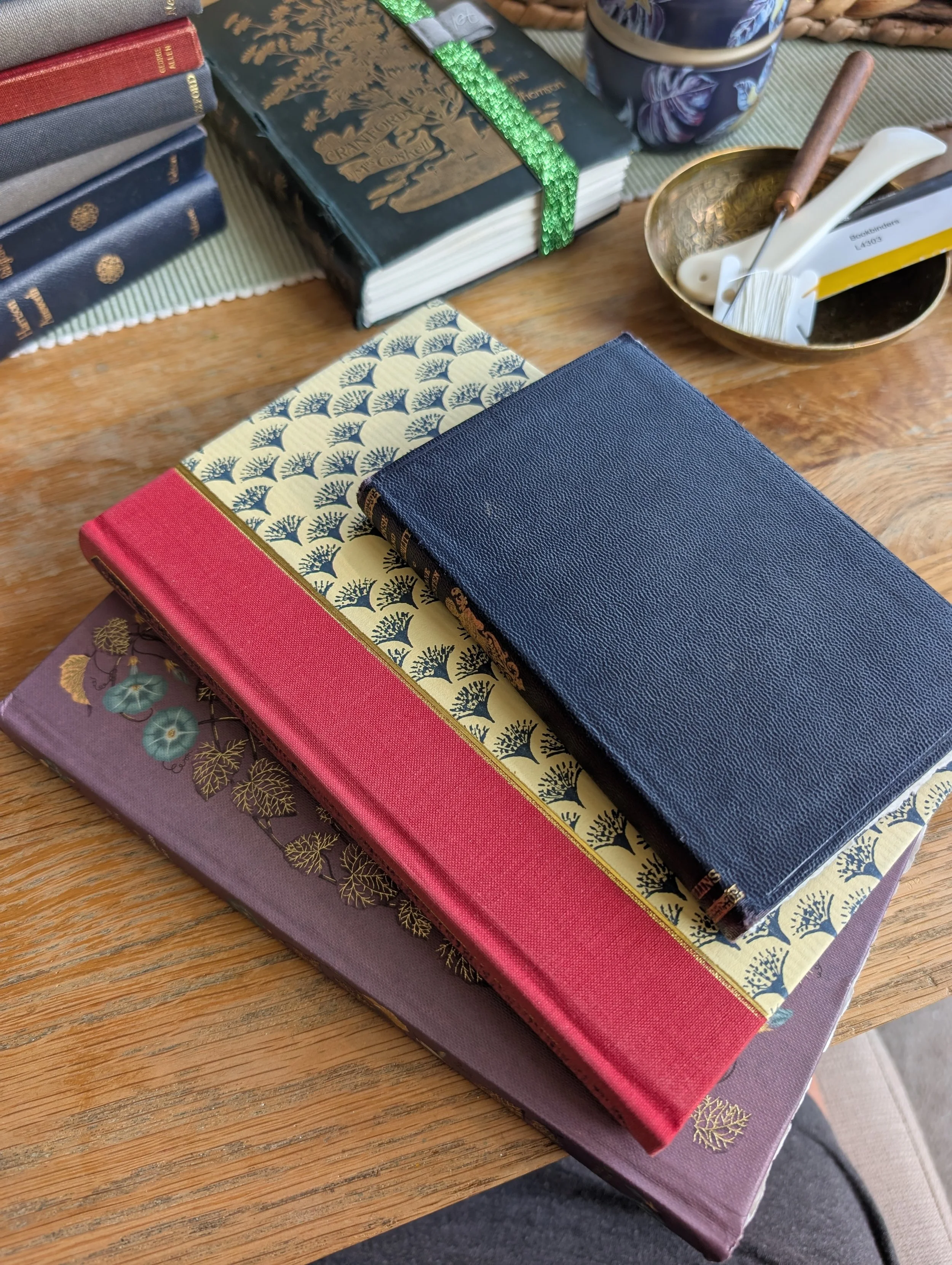 A collection of hardcover books with various decorative covers and colors, arranged on a wooden table with a placemat underneath, alongside a bowl containing patch tools and other small items.