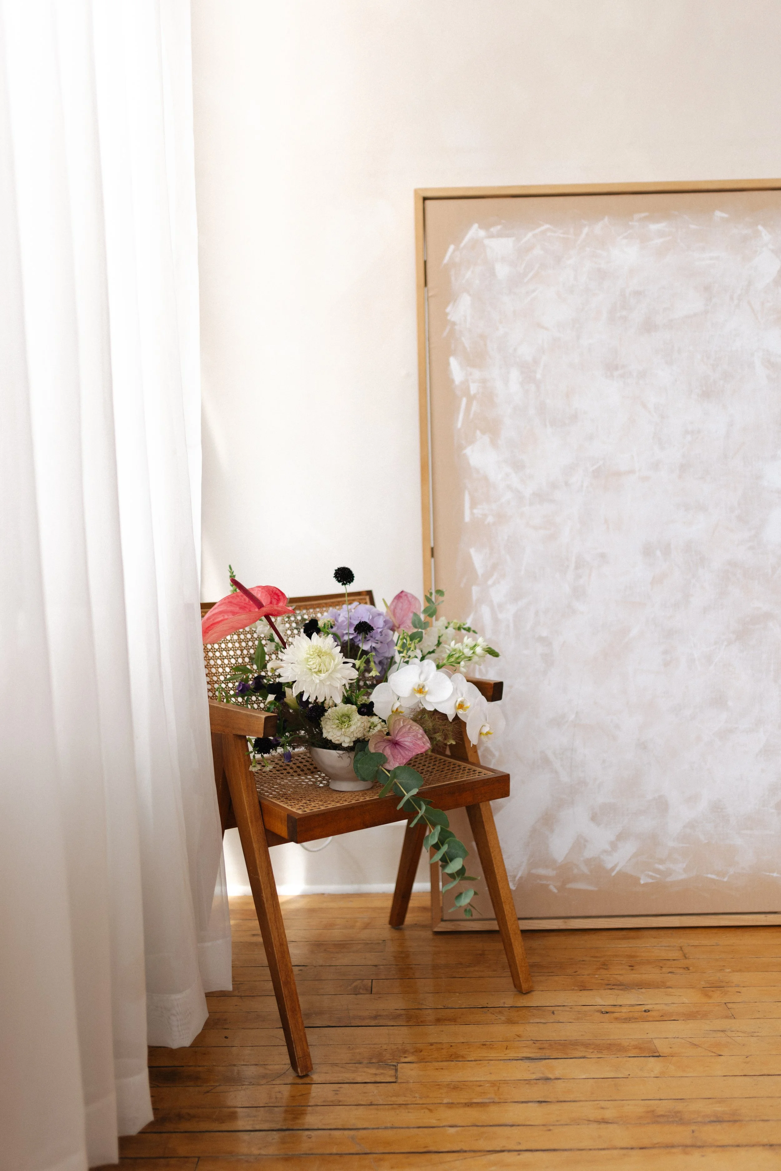 A wooden chair with a floral arrangement in a white bowl on it, positioned against a textured beige wall, with a white curtain partially covering the left side of the scene.