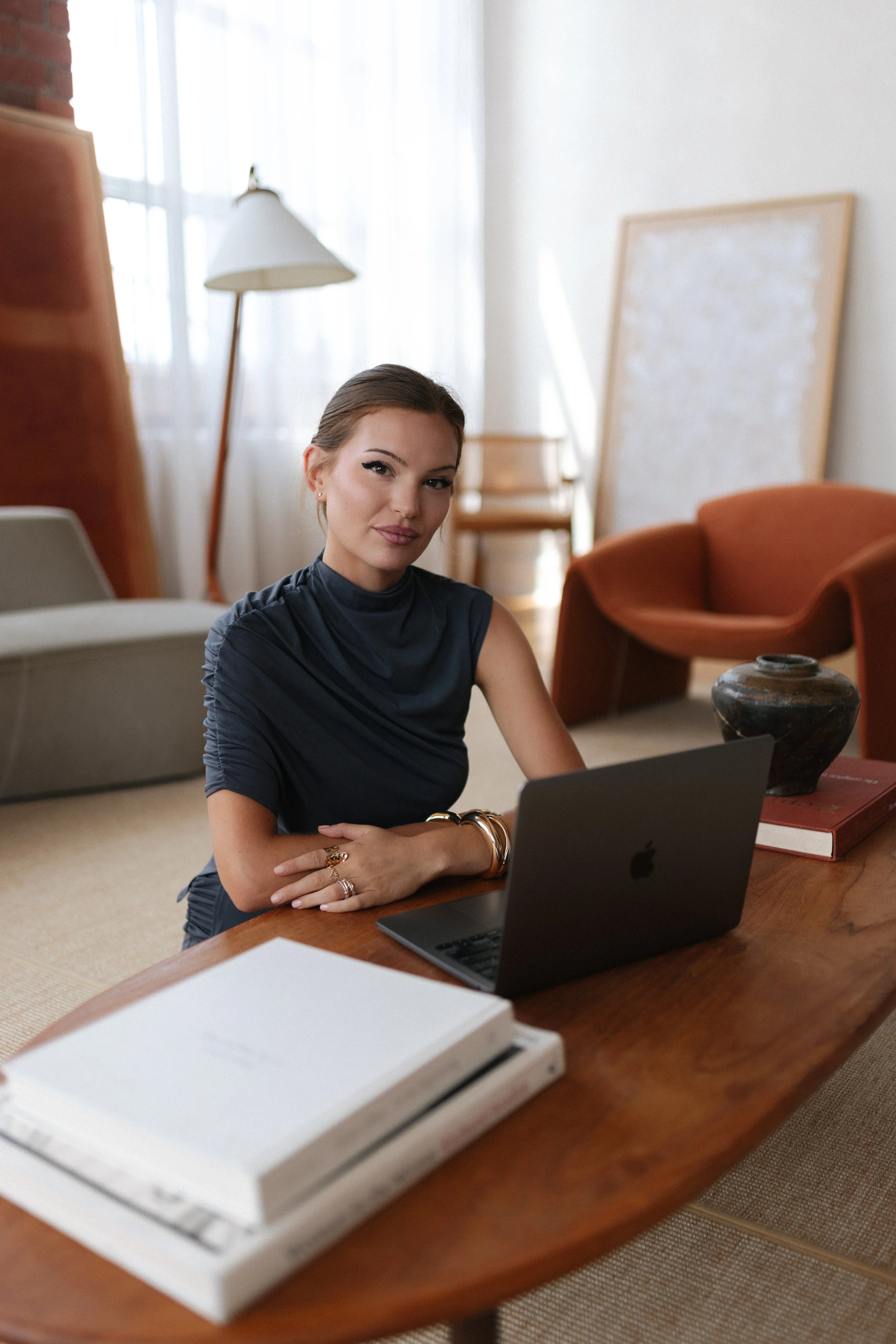 A woman with dark hair in a bun, wearing a dark sleeveless top, sitting at a wooden table with a laptop in front of her, in a room with white walls, a standing lamp, a chair, a picture frame, and a large decorative jar.