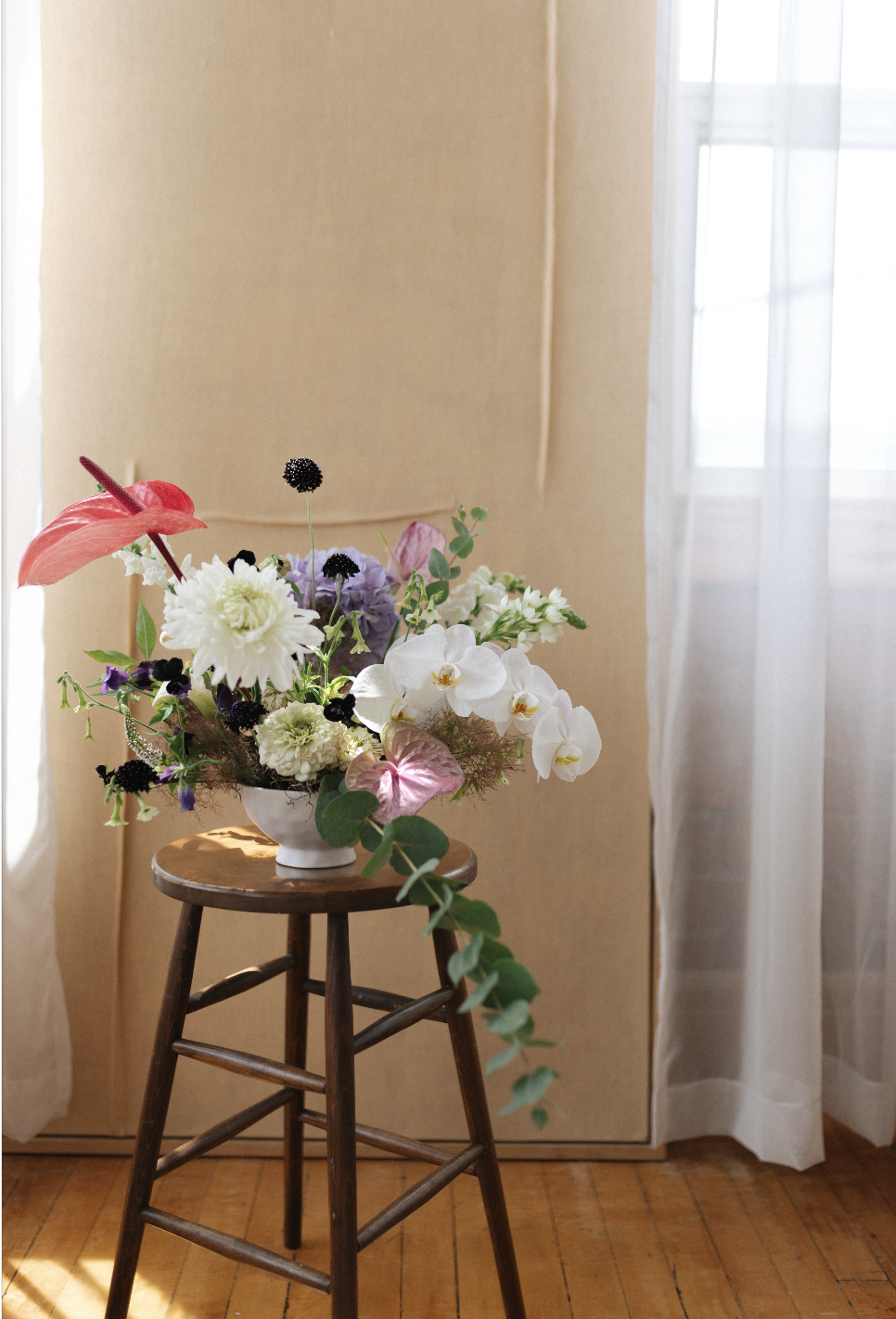 A white flower arrangement in a white bowl on a wooden stool, with various colorful flowers and greenery, in front of a beige wall and near a window with sheer curtains.