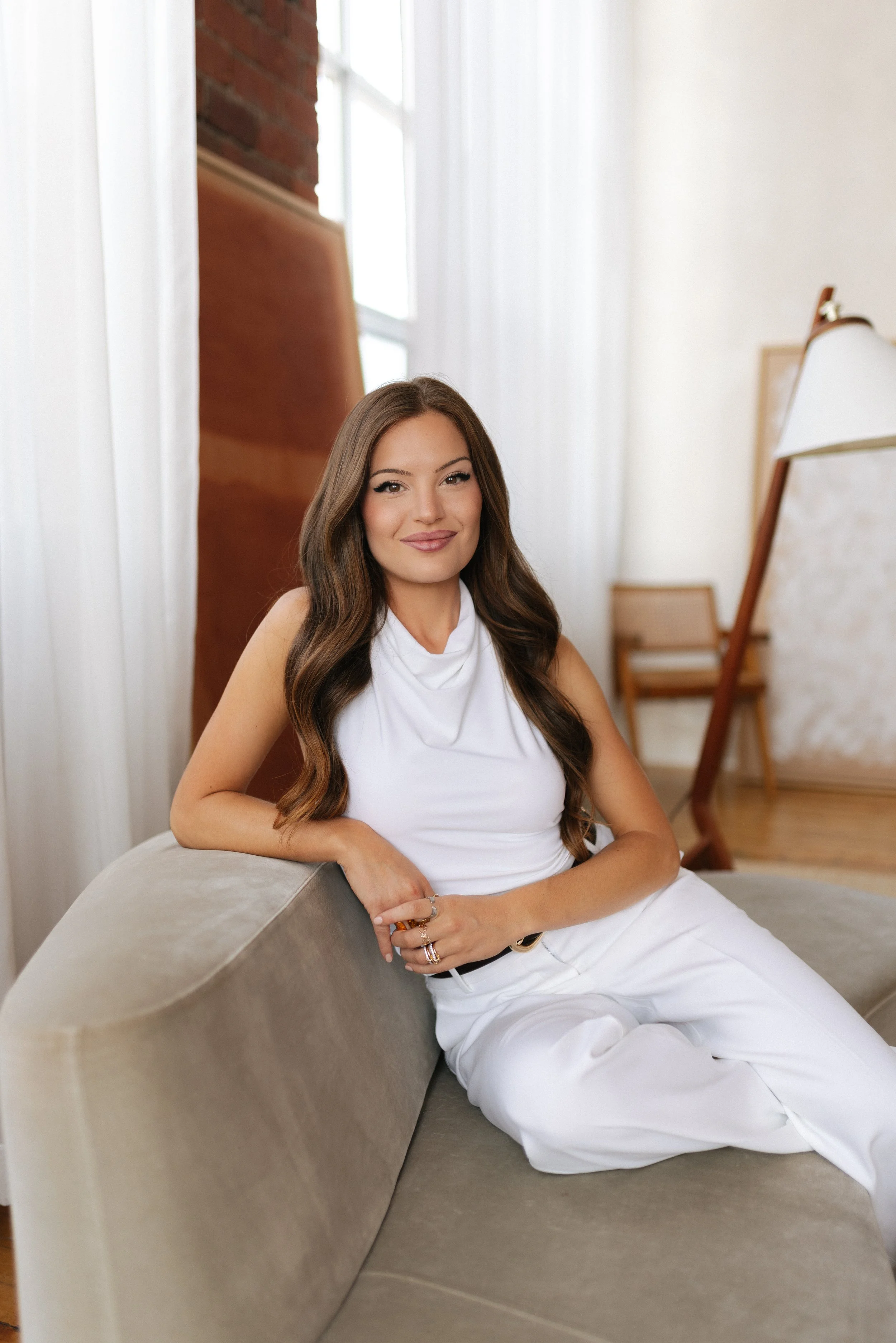 A young woman with long brown hair, wearing a white sleeveless top and white pants, sitting on a curved beige sofa in a bright, modern room with large windows and white curtains, smiling at the camera.