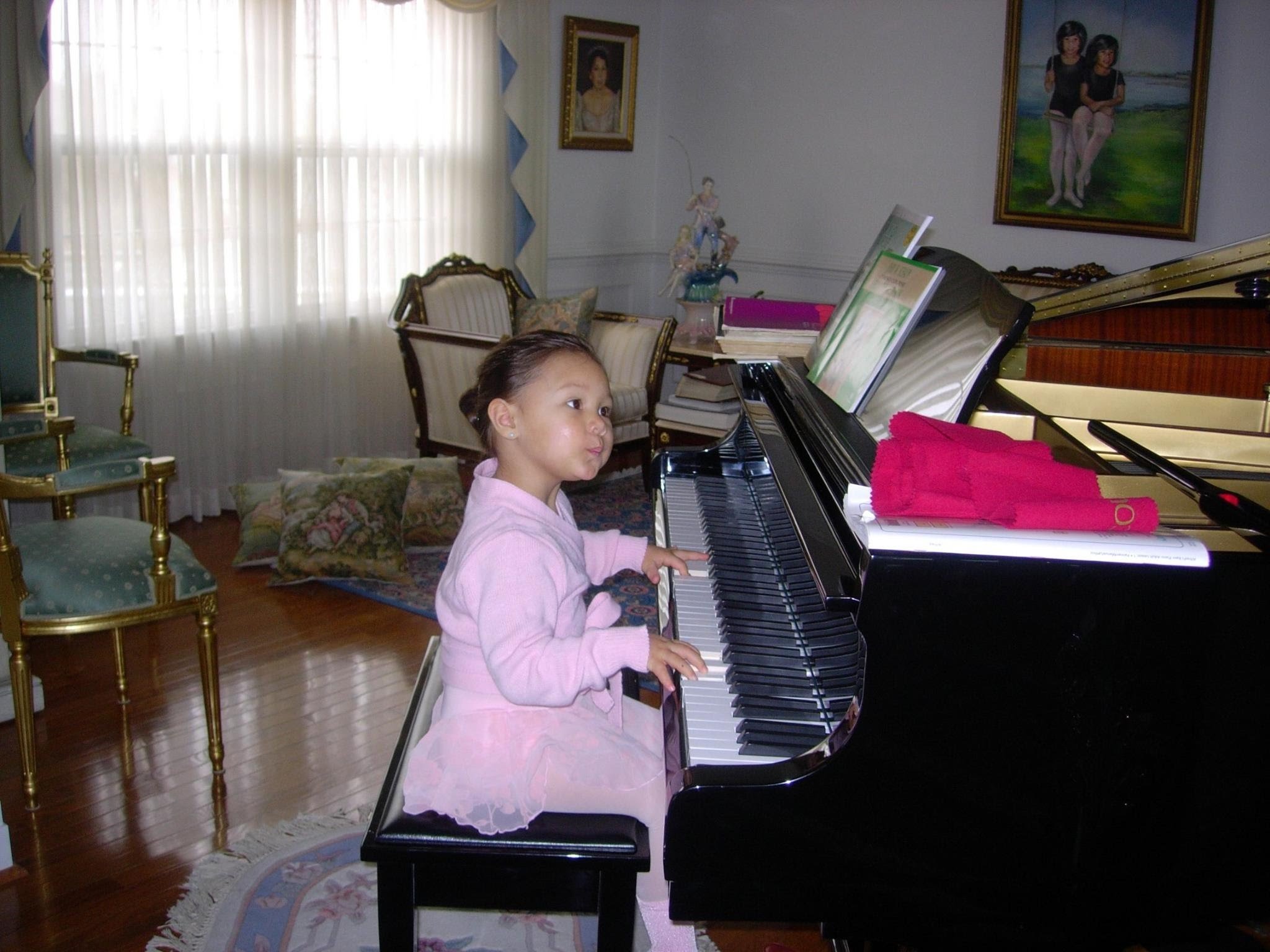 A young girl in a pink dress playing a grand piano in a decorated living room with artwork on the walls and a window with curtains.