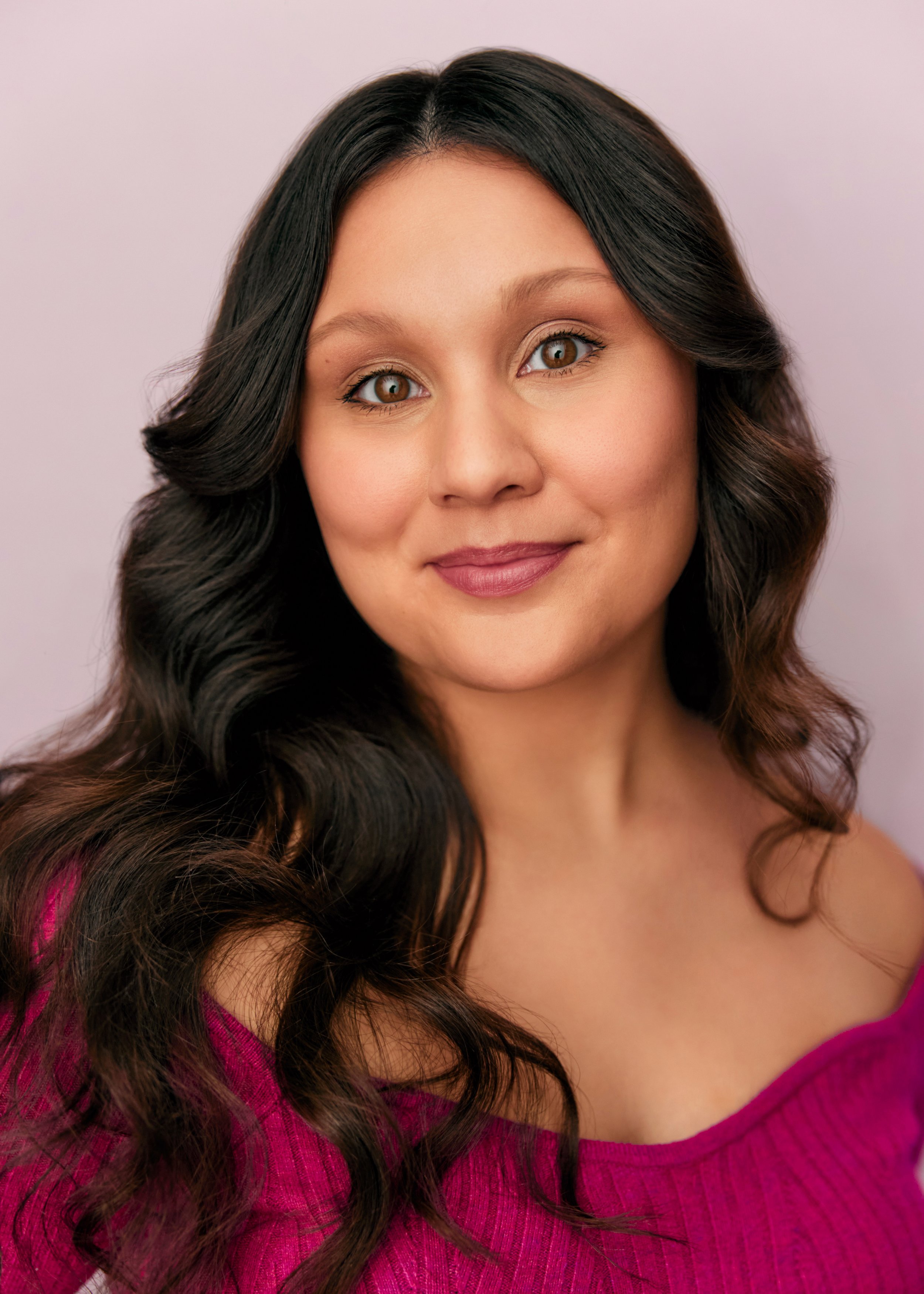 A young woman with long, dark wavy hair and light brown eyes smiling in front of a plain, light pink background. She is wearing a magenta sweater.