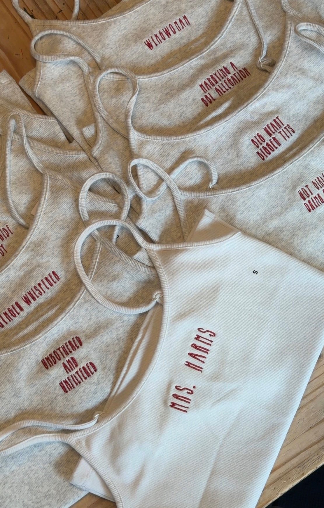 Multiple gray and white tank tops with red text that read phrases such as 'Unbothered and Unfiltered' and 'Wingwoman' laid out on a wooden surface.