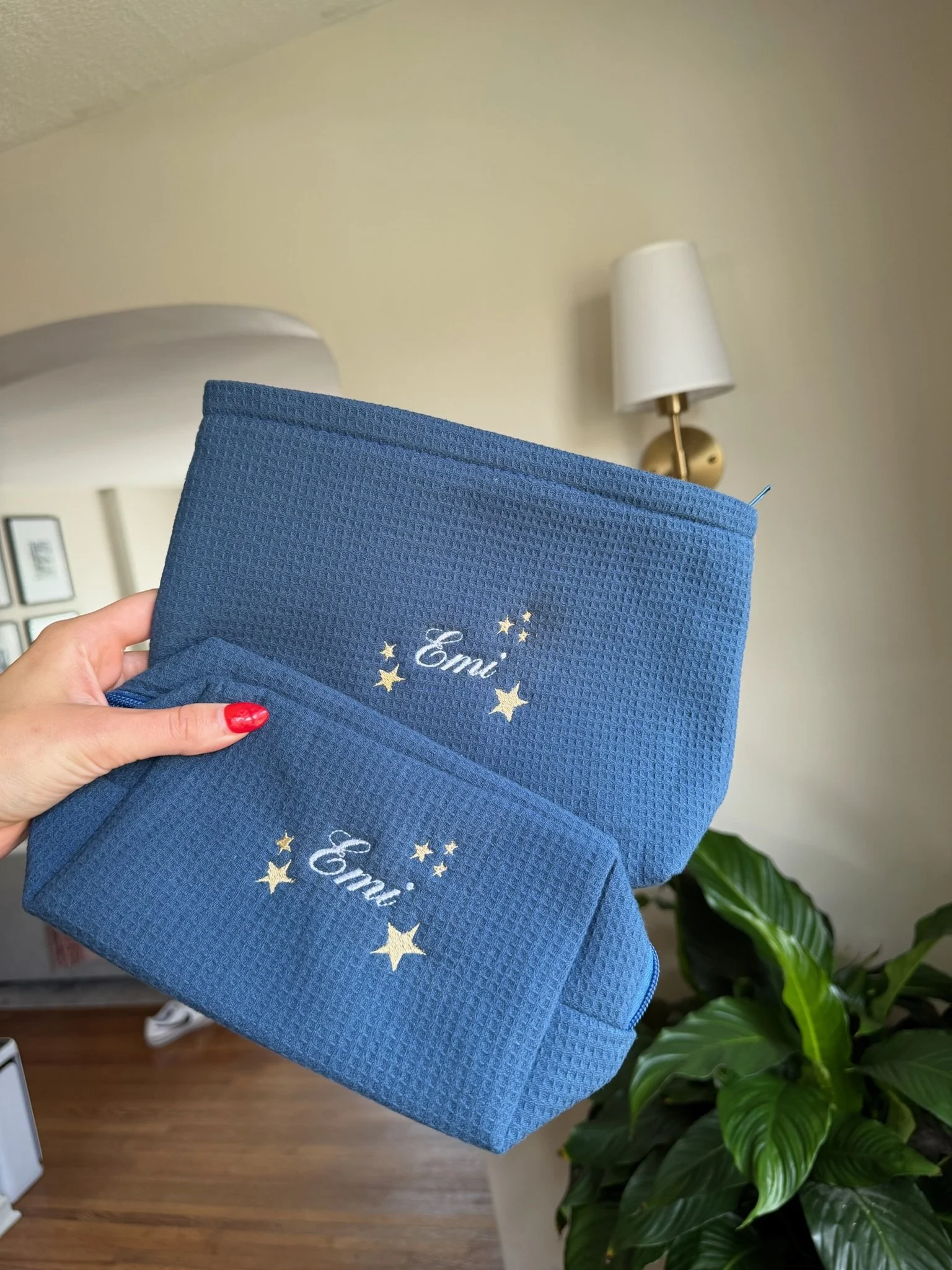 A person with red nail polish holding two blue fabric items embroidered with the name Emi and gold stars on a wall with a white lamp.