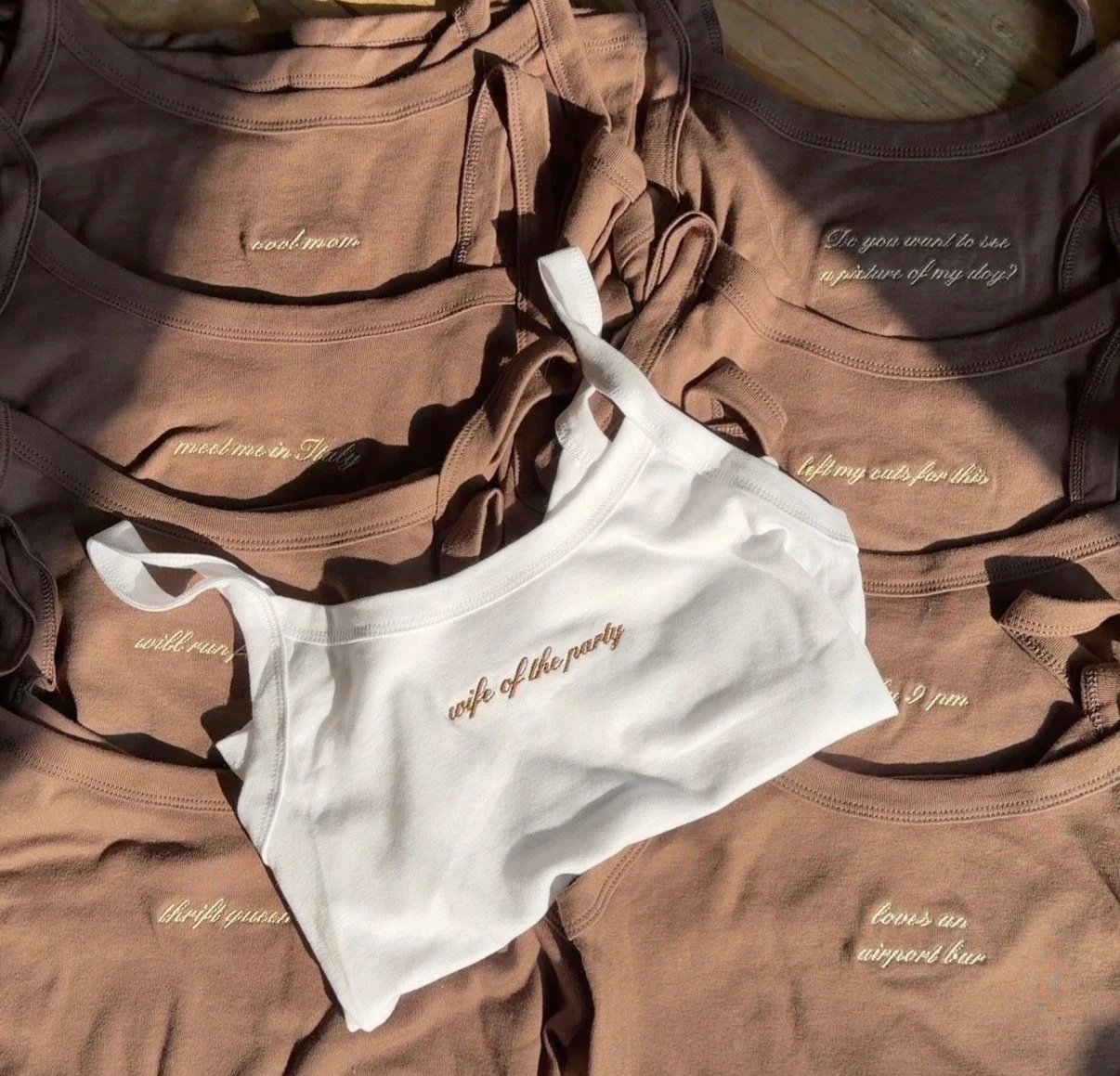 Multiple beige and white t-shirts with embroidered text, laid out flat, with one white t-shirt on top that has the words "wife of the party" embroidered in gold.