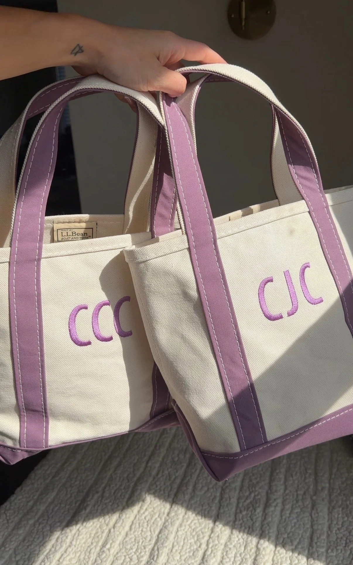 Two beige tote bags with purple straps and accents, embroidered with the letter 'C' or 'CC' in purple, held by a person's hand with a small tattoo on the wrist, placed on a light-colored surface with sunlight casting shadows.