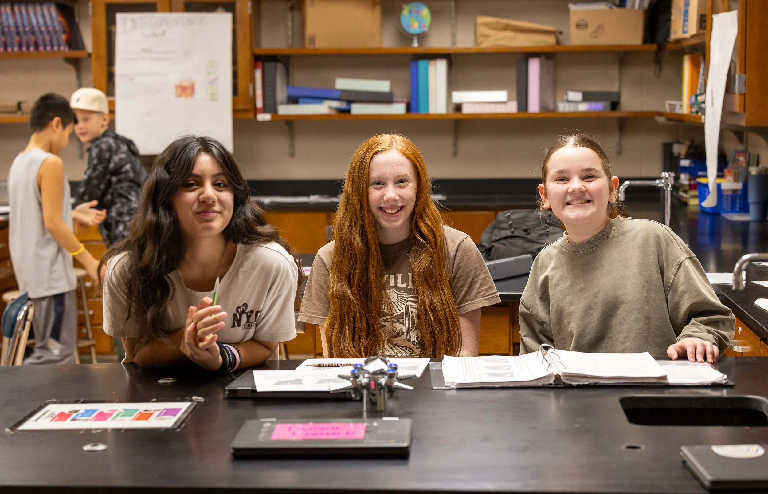 Two smiling high school students sitting at a science classroom table with open notebooks and science supplies.