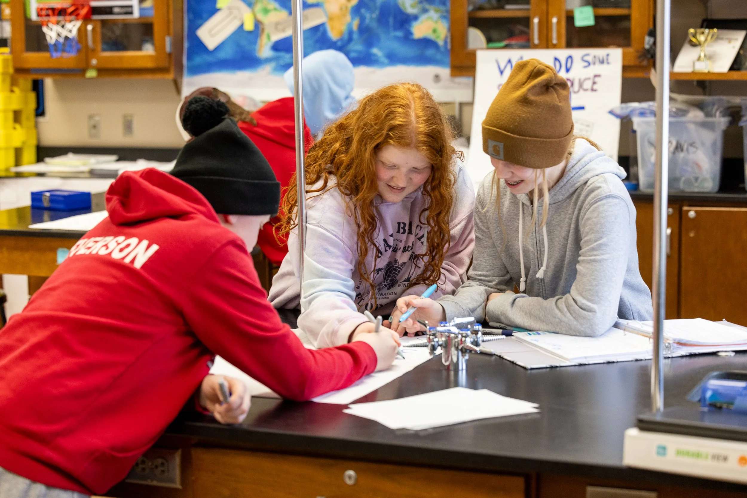 Three students working together at a science table in a classroom, with a world map and educational posters in the background.