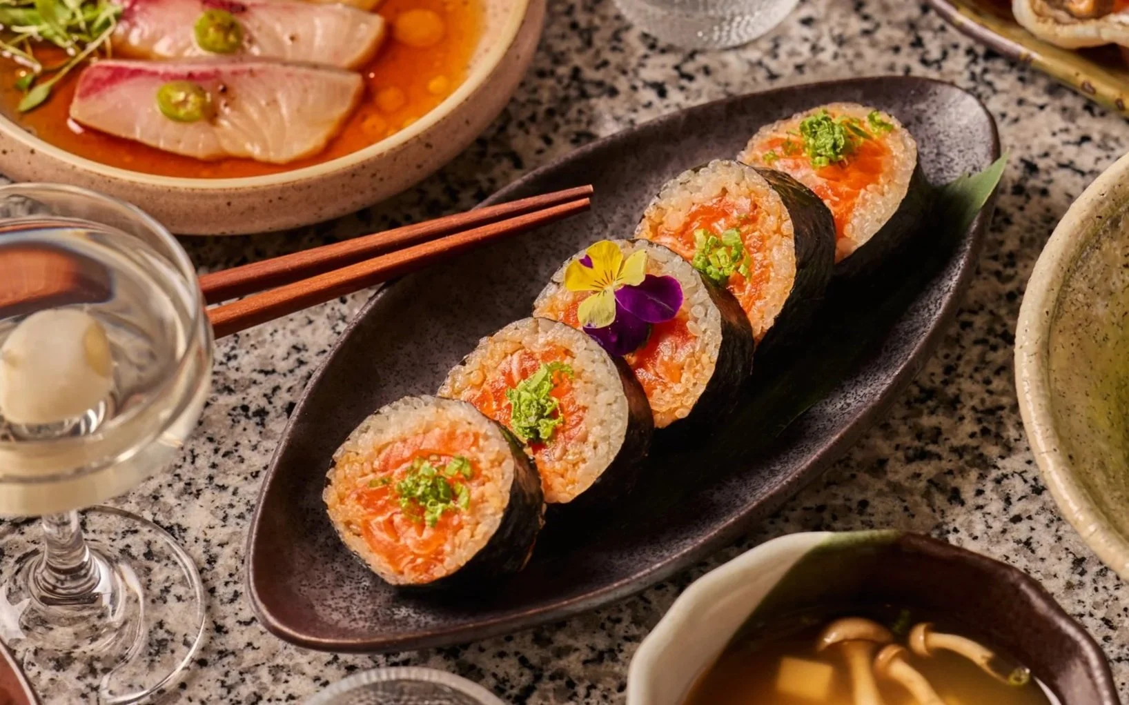 Sushi rolls with rice, salmon, seaweed wrap, garnished with flowers, green herbs, on a black plate; other Japanese dishes and drink around.