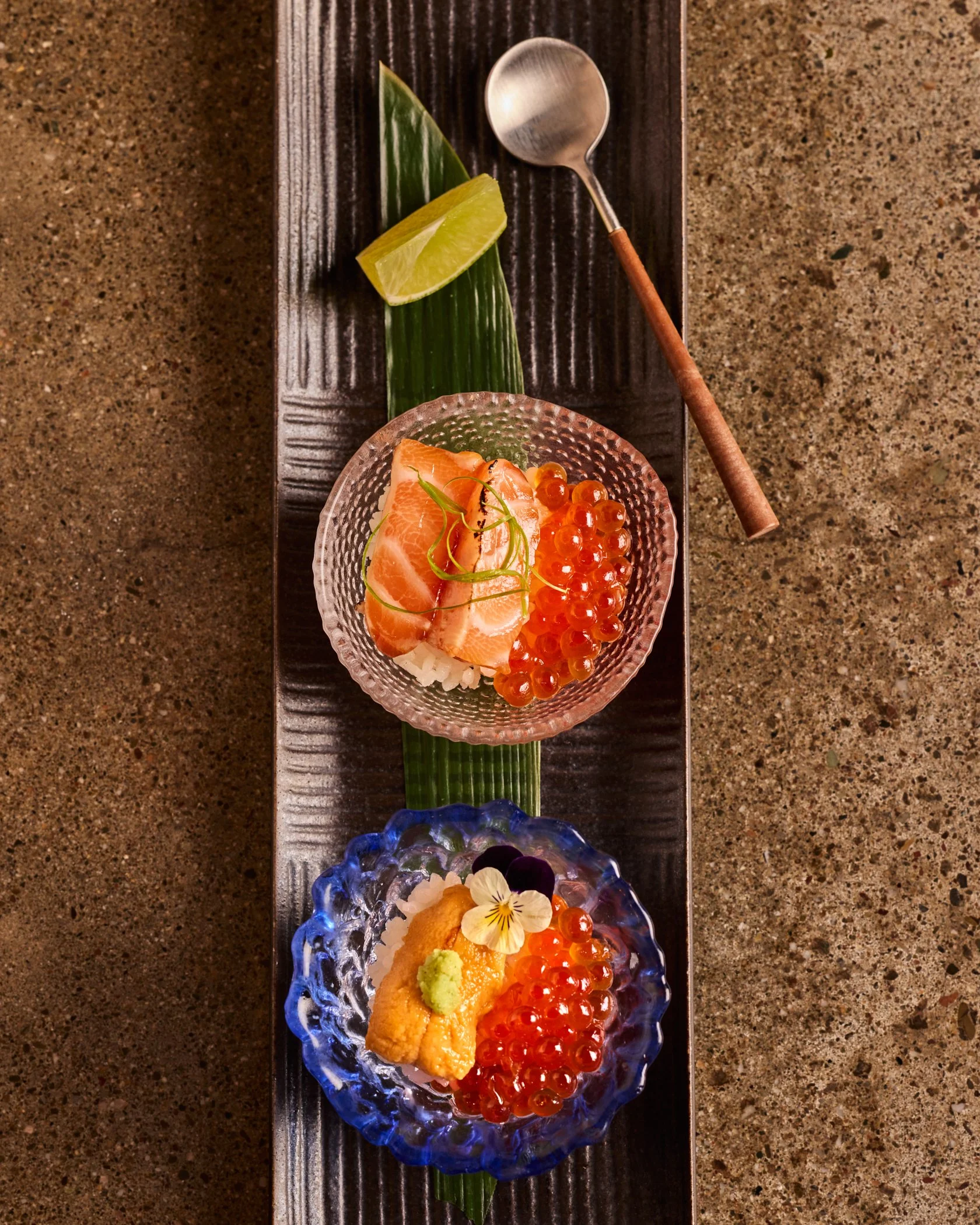 Plate of Japanese cuisine topped with two small bowl, a spoon and a lime wedge. The top bowl has sushi rice topped with ikura and uni. The bottom bowl has sushi rice topped with ikura and a small piece of salmon.