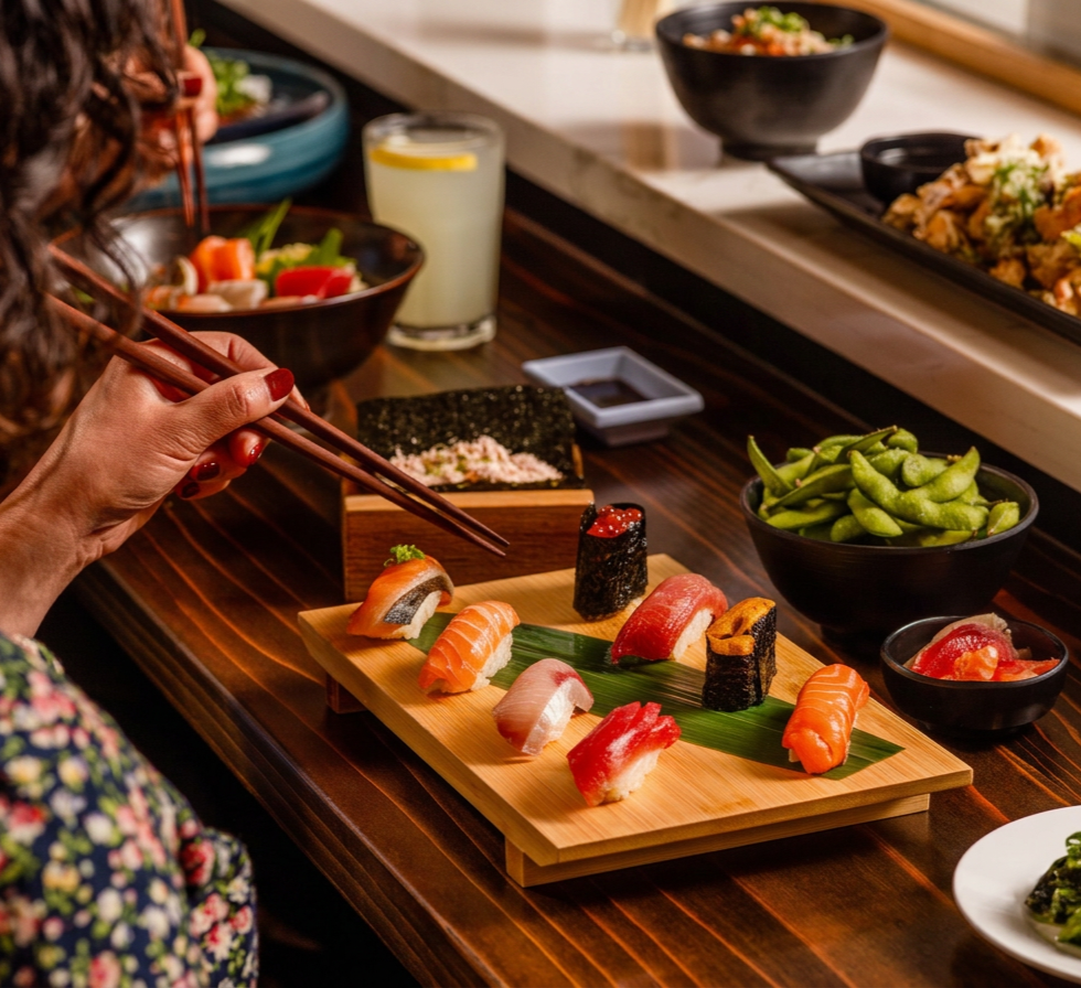 Person using chopsticks to pick up sushi on a wooden serving board, surrounded by bowls of edamame, sauces, and other Japanese dishes in a restaurant setting.