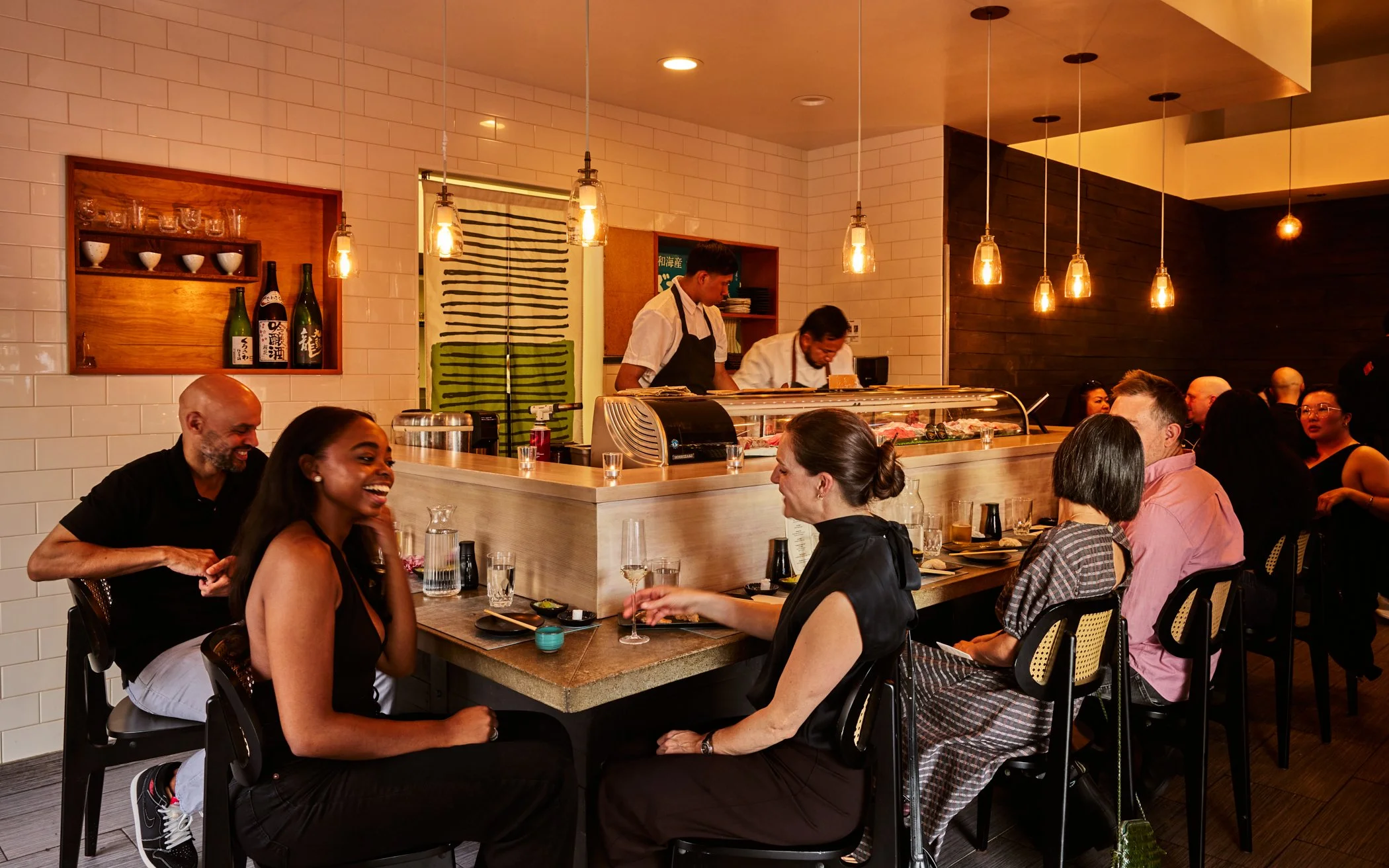 A lively sushi restaurant with patrons seated at tables, some smiling and engaging in conversation, while chefs prepare sushi behind the counter. The interior features white tiled walls, warm lighting, and wooden accents.