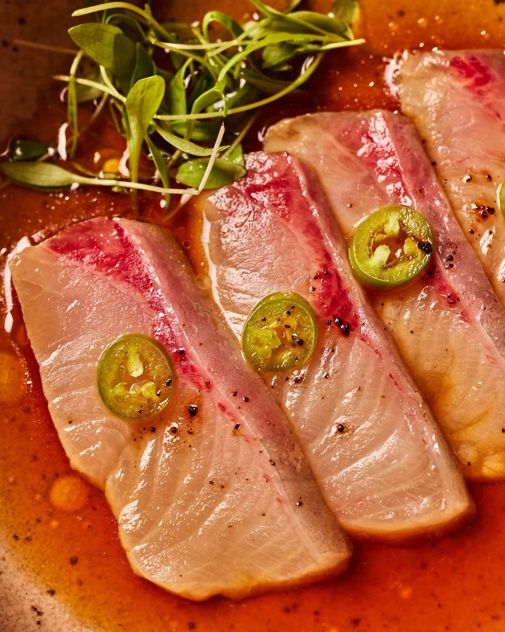Close-up of hamachi carpaccio with slices of serrano and fresh green herbs on top, sitting in a ponzu sauce.