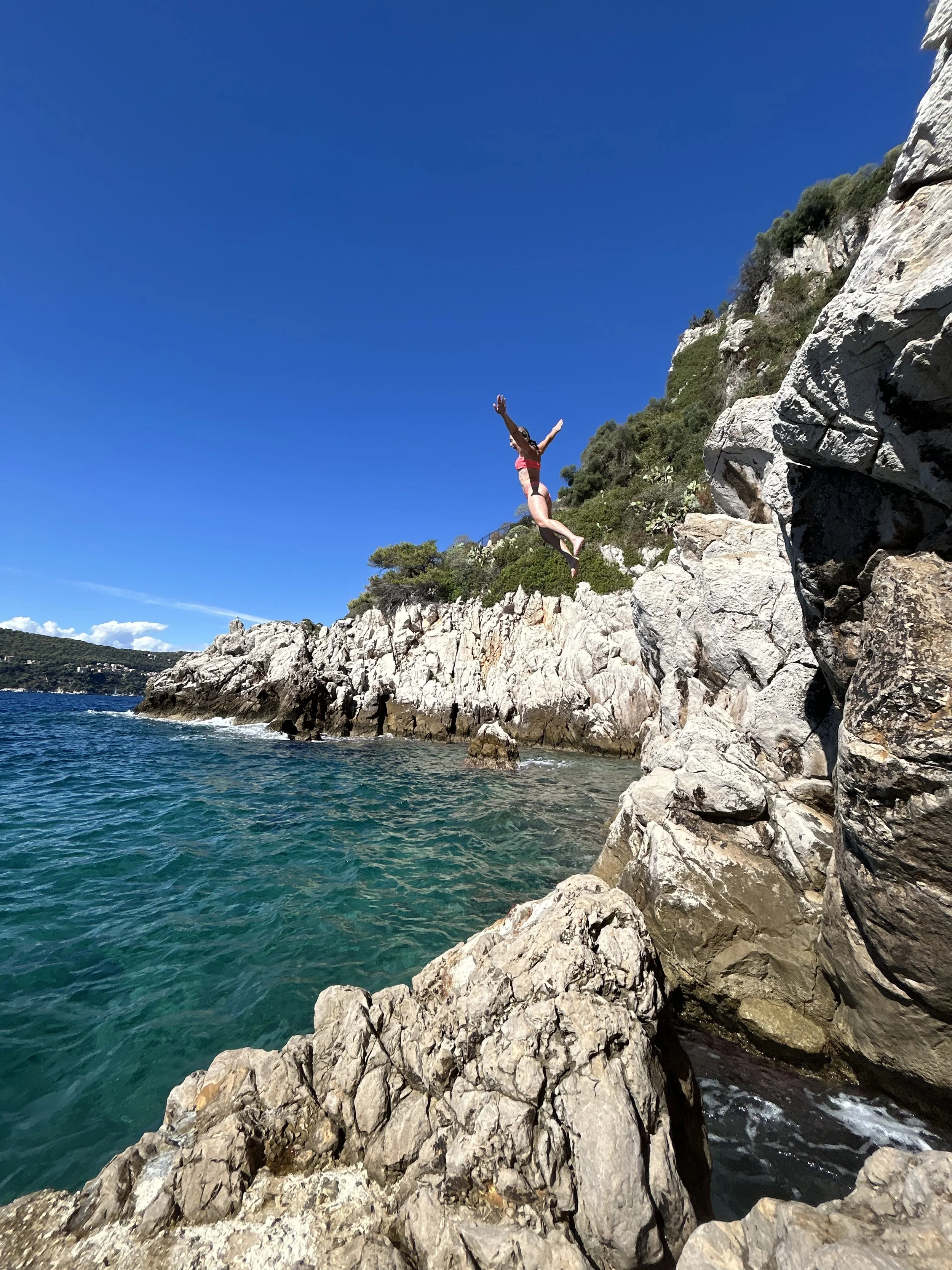 A person jumping off a rocky cliff into clear blue water, with a bright blue sky and green hills in the background.