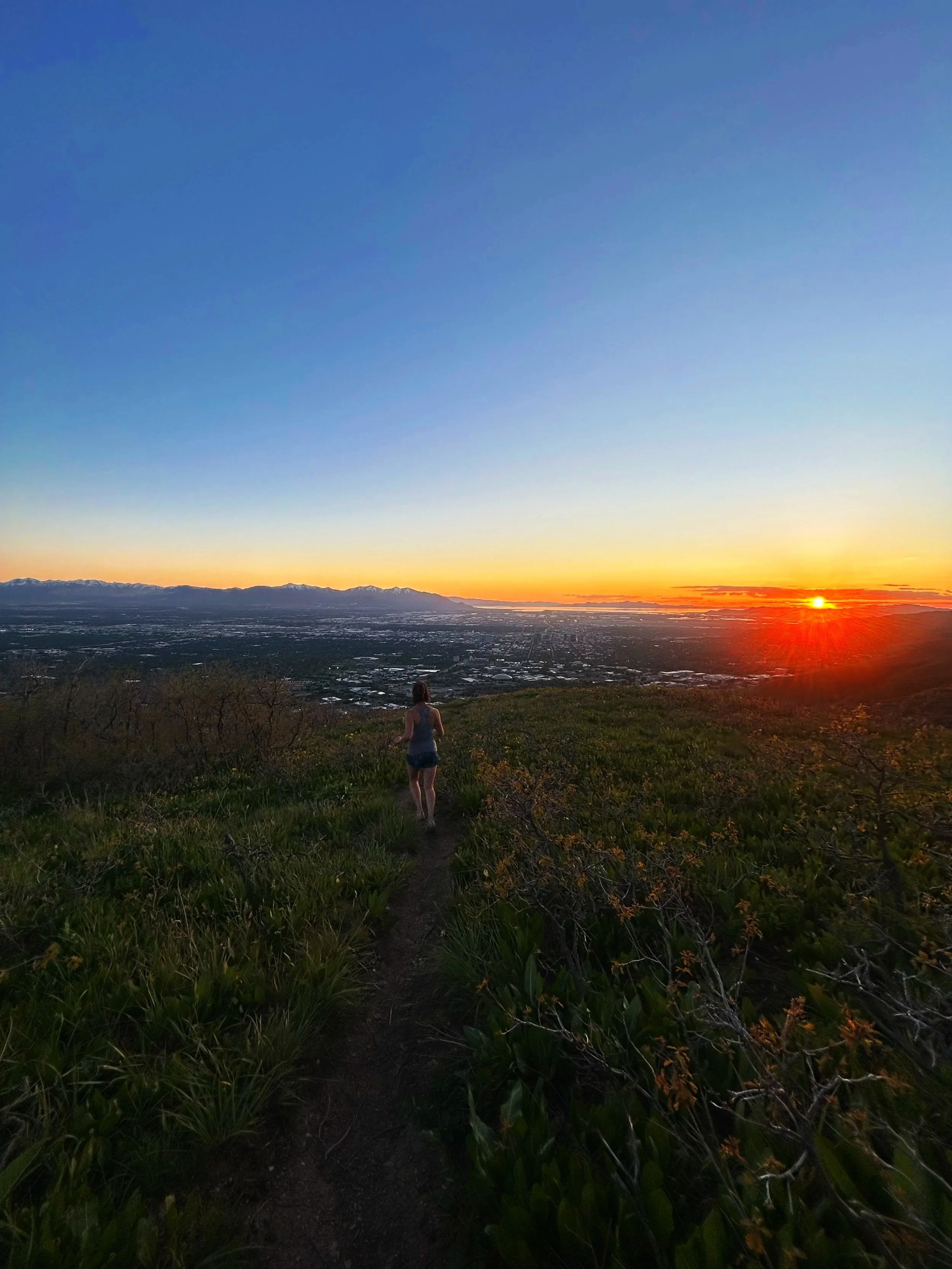 A person walking on a trail through a grassy hillside at sunset, with mountains in the distance and the sun setting on the horizon.