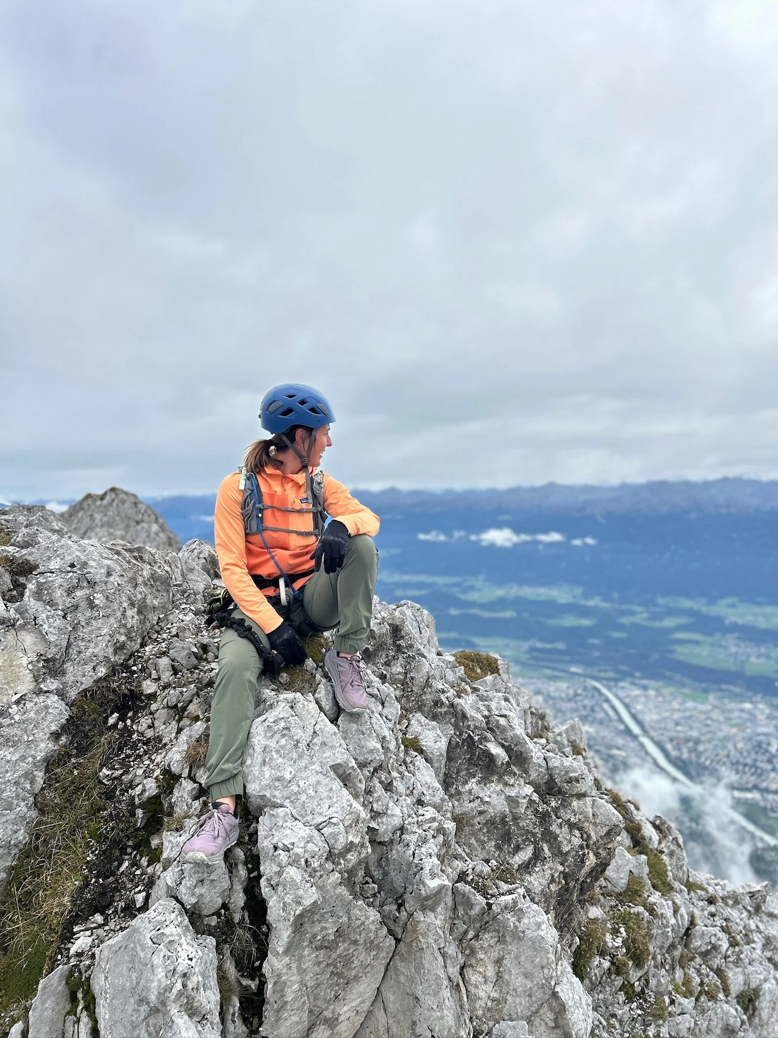A woman wearing a blue helmet, orange jacket, and gray pants sitting on rocks on a mountain with a scenic view of valleys, rivers, and distant mountains under cloudy skies.