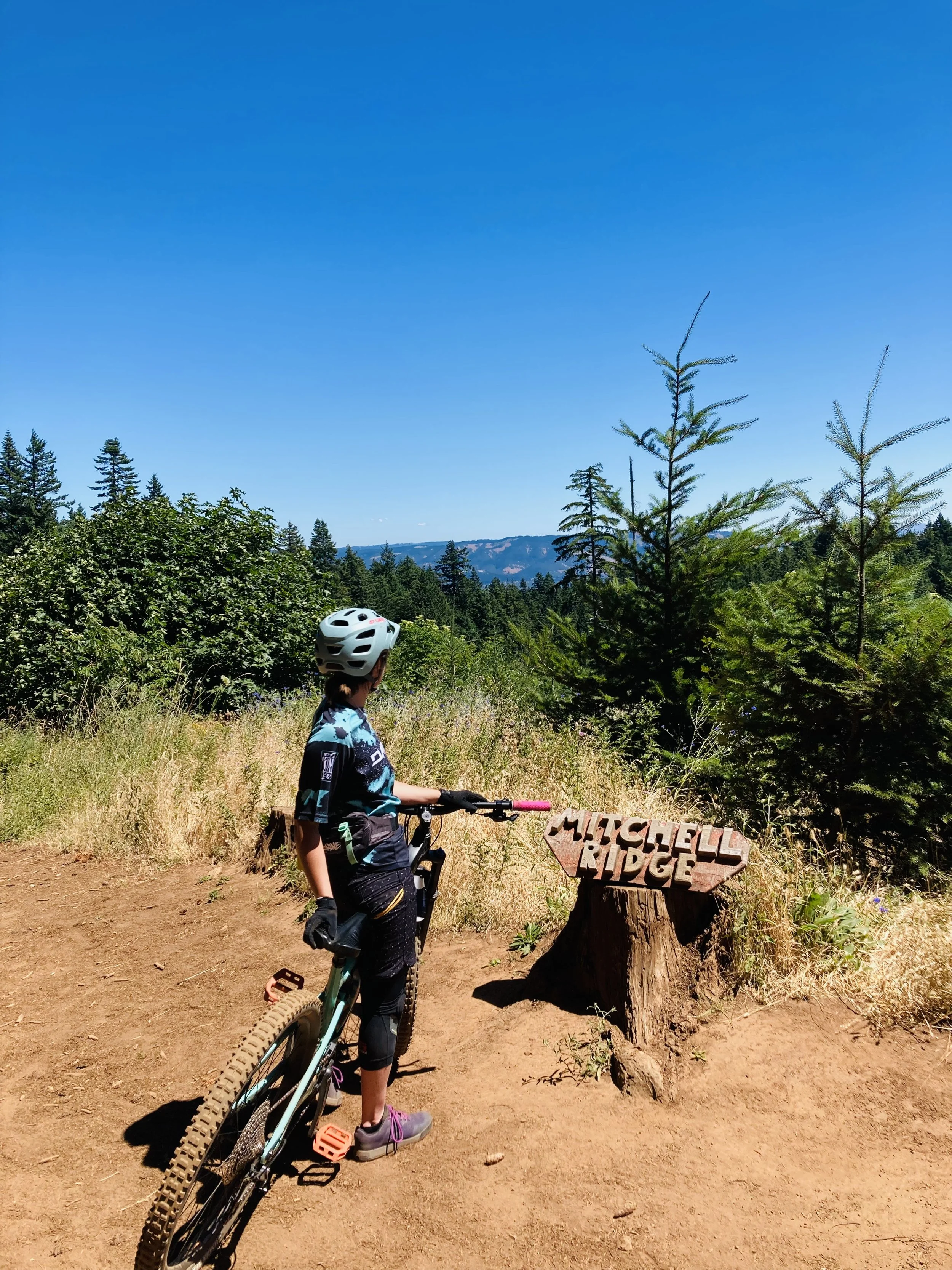 A young girl with a mountain bike standing on a dirt trail in a forested area with mountains in the background. She is wearing a helmet, gloves, and athletic clothes, and is looking toward a wooden sign that reads 'MITCHELL RIDGE.'