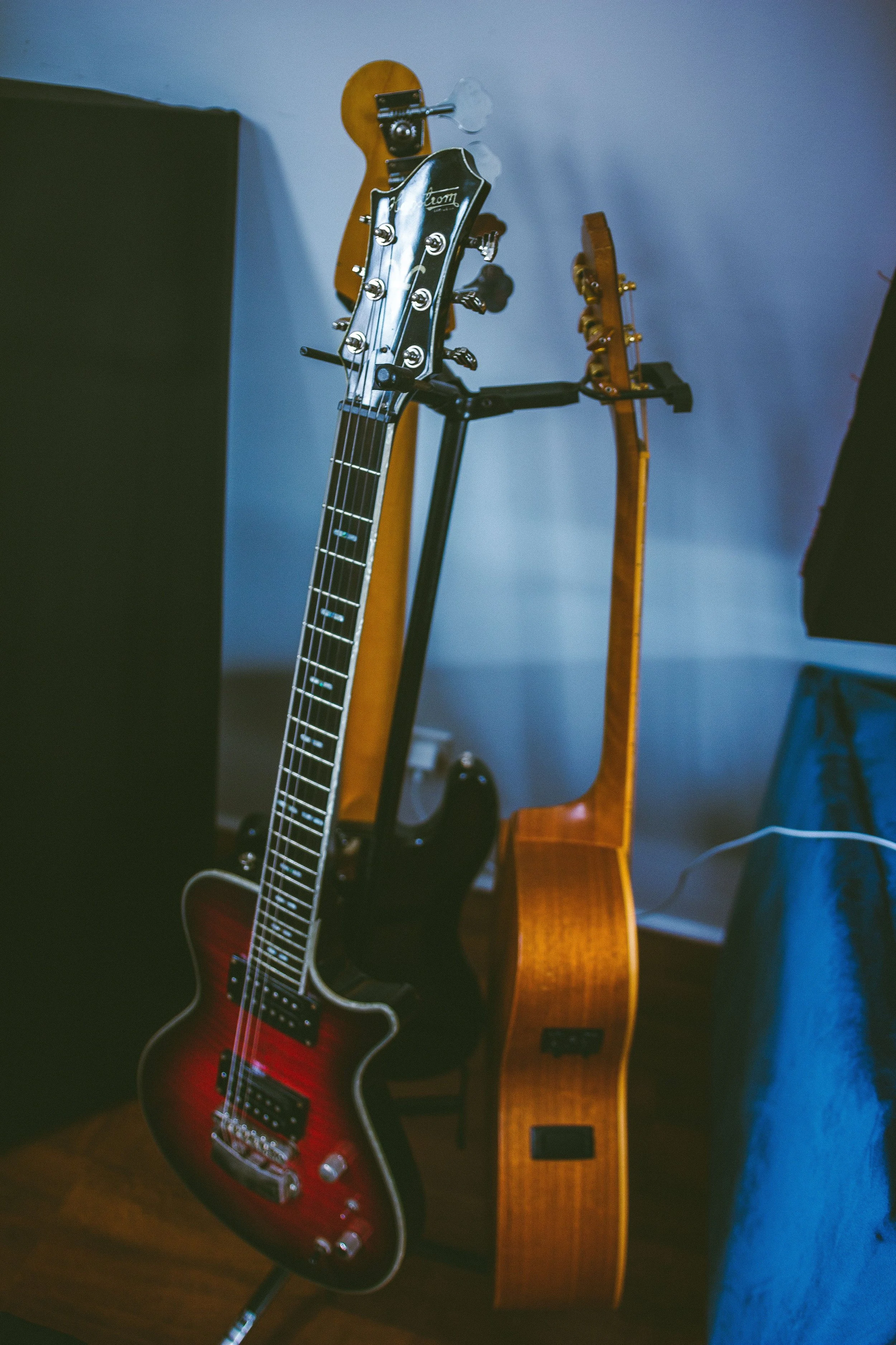 Two guitars, one red electric with black accents and one natural wood acoustic, standing on a guitar rack in a room with a black speaker and blue walls.