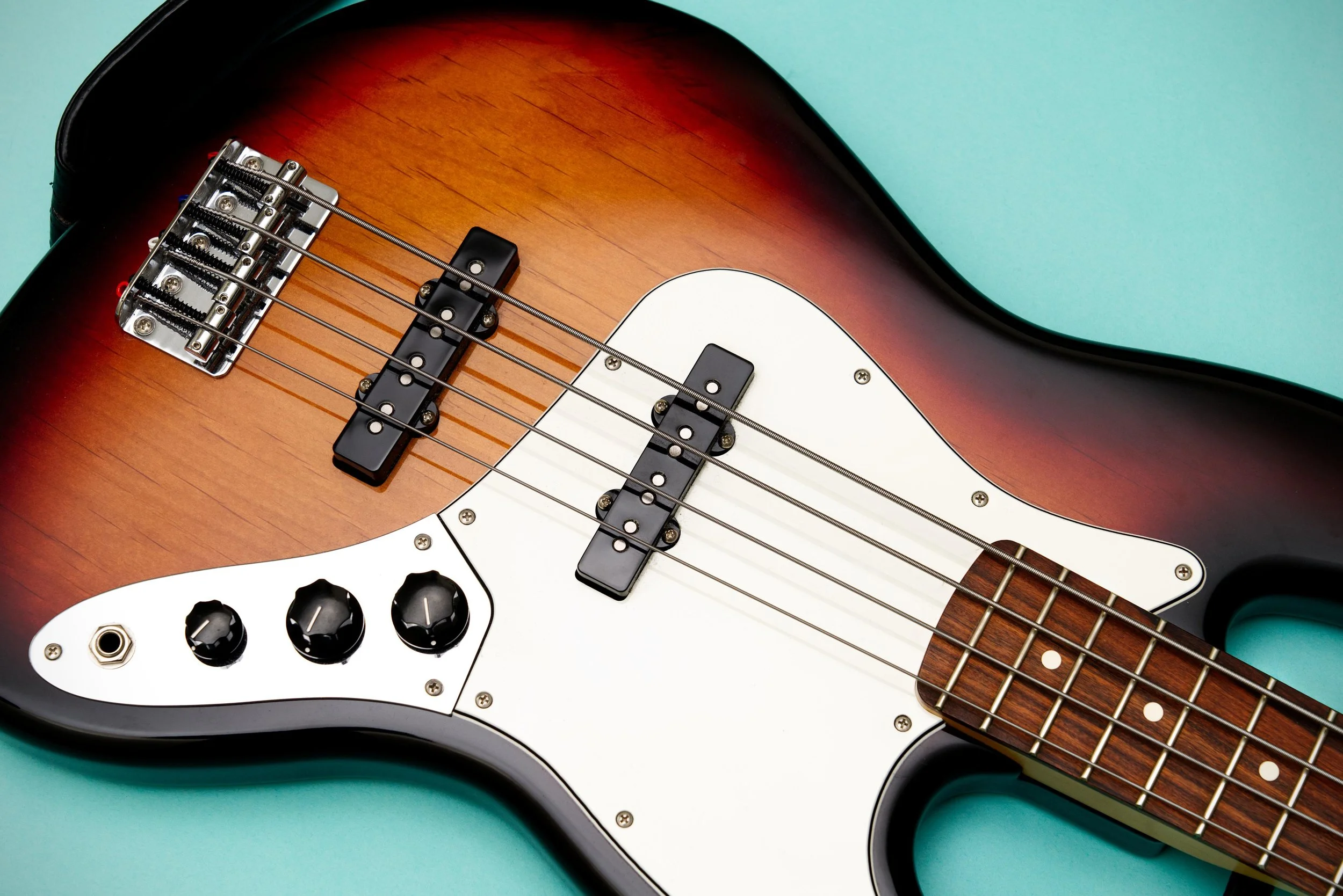 Close-up of an electric bass guitar with a sunburst finish, white pickguard, black pickups, and control knobs, on a teal background.
