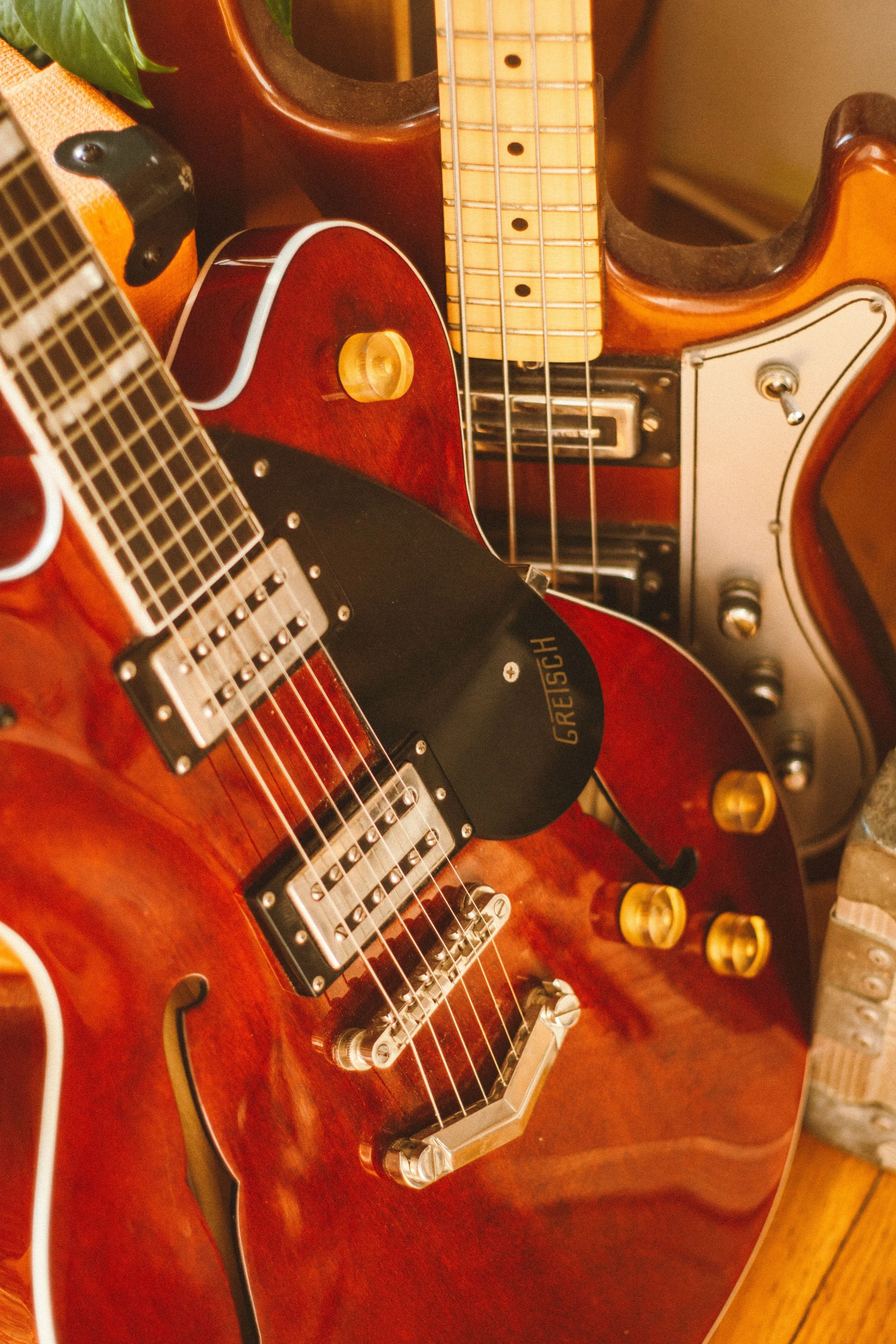 Close-up of a collection of musical instruments including an electric guitar with a reddish-brown body, a vintage acoustic guitar, and a red electric bass guitar with a light-colored neck, all placed against a warm-colored background.
