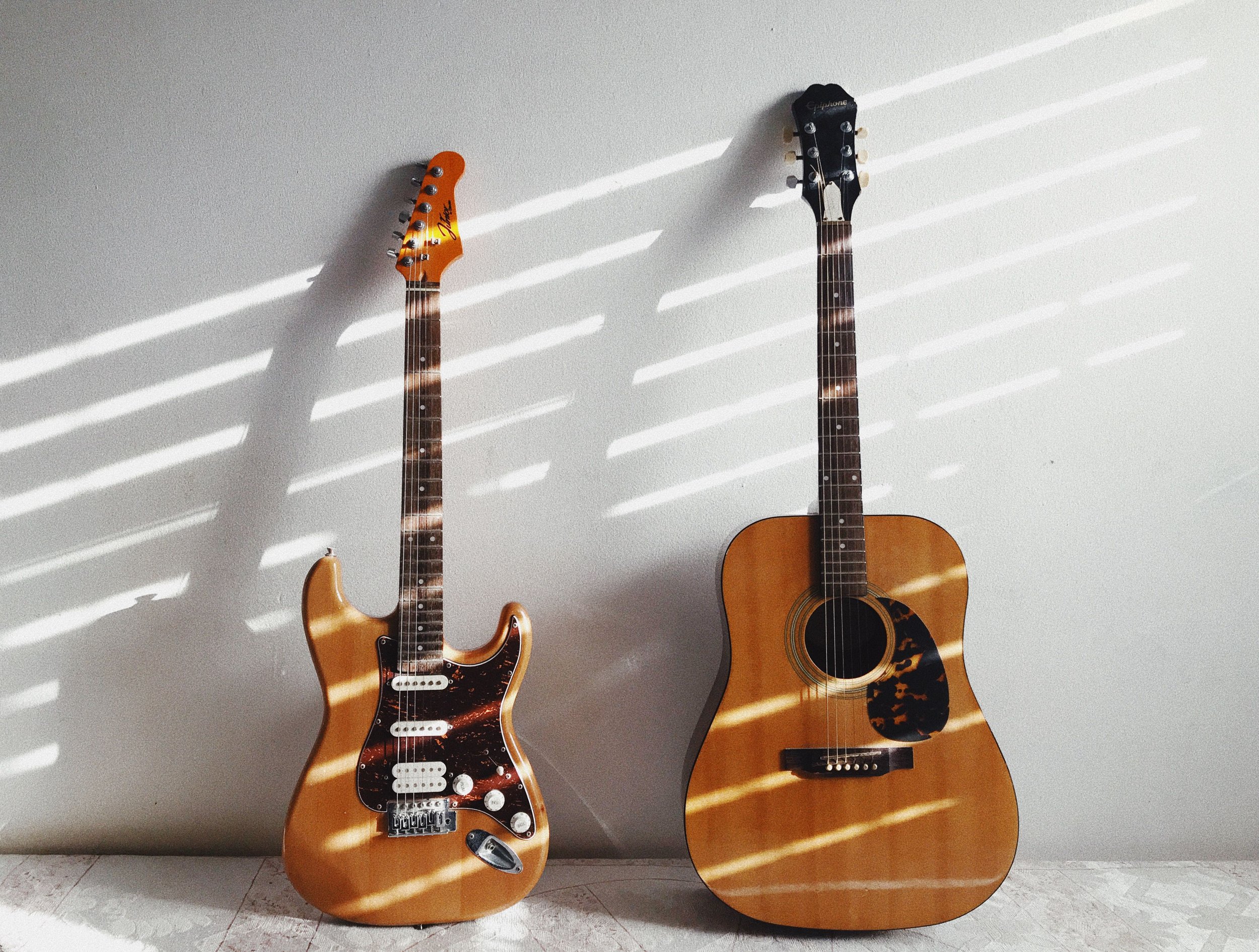 Two guitars, one electric with a wood finish and tortoiseshell pickguard, and one acoustic with a natural wood finish, leaning against a wall with light hitting it through blinds creating striped shadows.