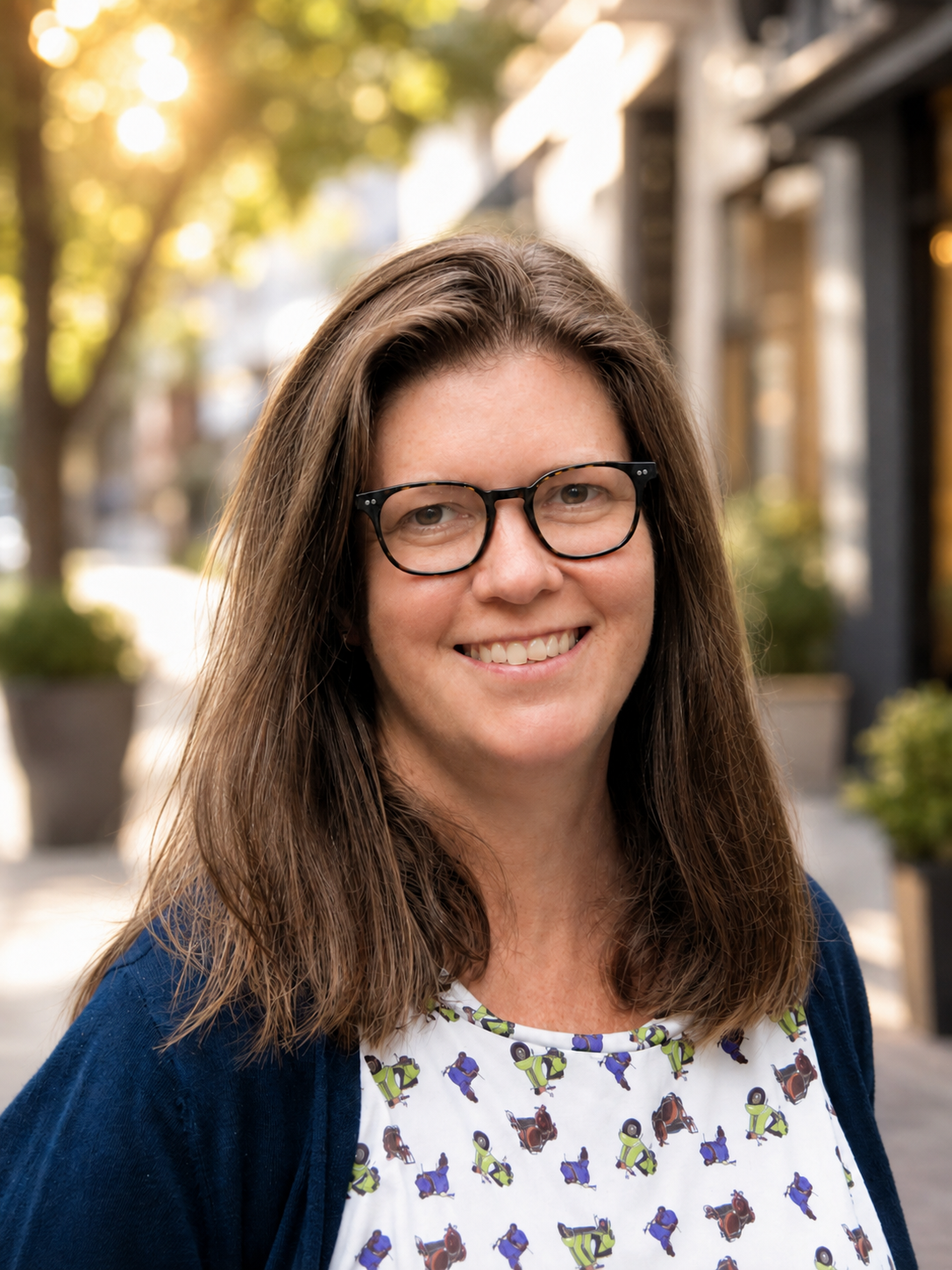 A woman with shoulder-length brown hair wearing glasses, a printed shirt, and a dark blue cardigan outdoors on a sunny day, smiling.