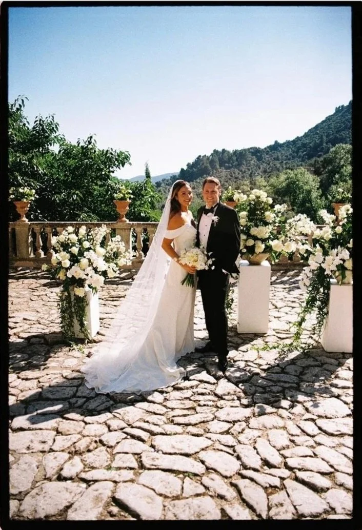 A bride and groom standing outdoors on a stone patio, surrounded by white floral arrangements, with green trees and hills in the background, during their wedding celebration.