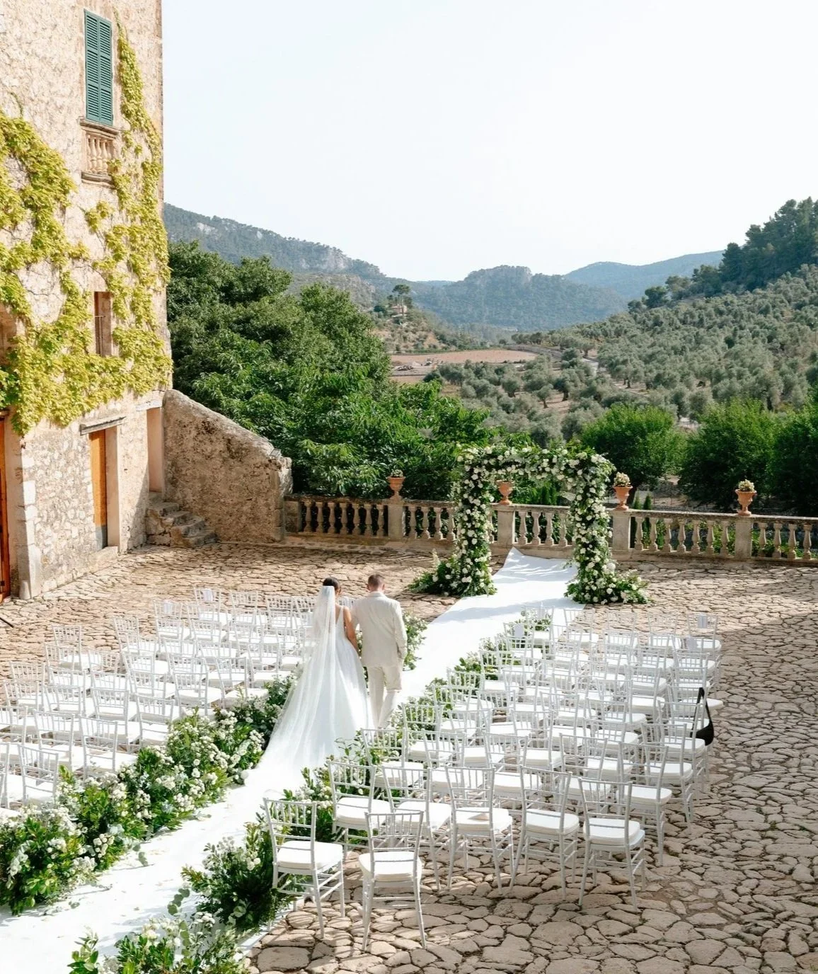 A bride and groom walking together down an outdoor wedding aisle decorated with white flowers, with rows of white chairs on either side, set against a scenic backdrop of rolling hills and greenery.