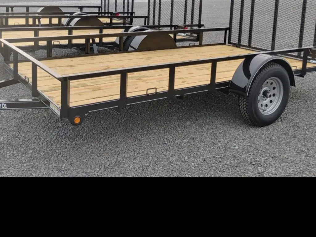 A flat utility trailer with a black metal frame, wooden floor, and single axle with a wheel on each side, parked on a gravel surface.