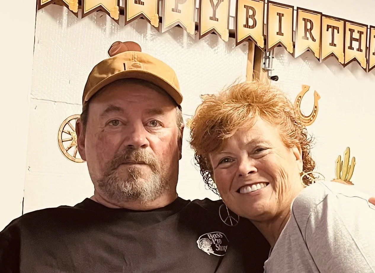 A man and a woman smiling together at a birthday celebration. The man has a beard, is wearing a brown cap and a black T-shirt with a logo. The woman has curly red hair and is wearing a light gray shirt and earrings. There is a birthday banner in the background with gold and black lettering and decorations, including horseshoes and cacti.