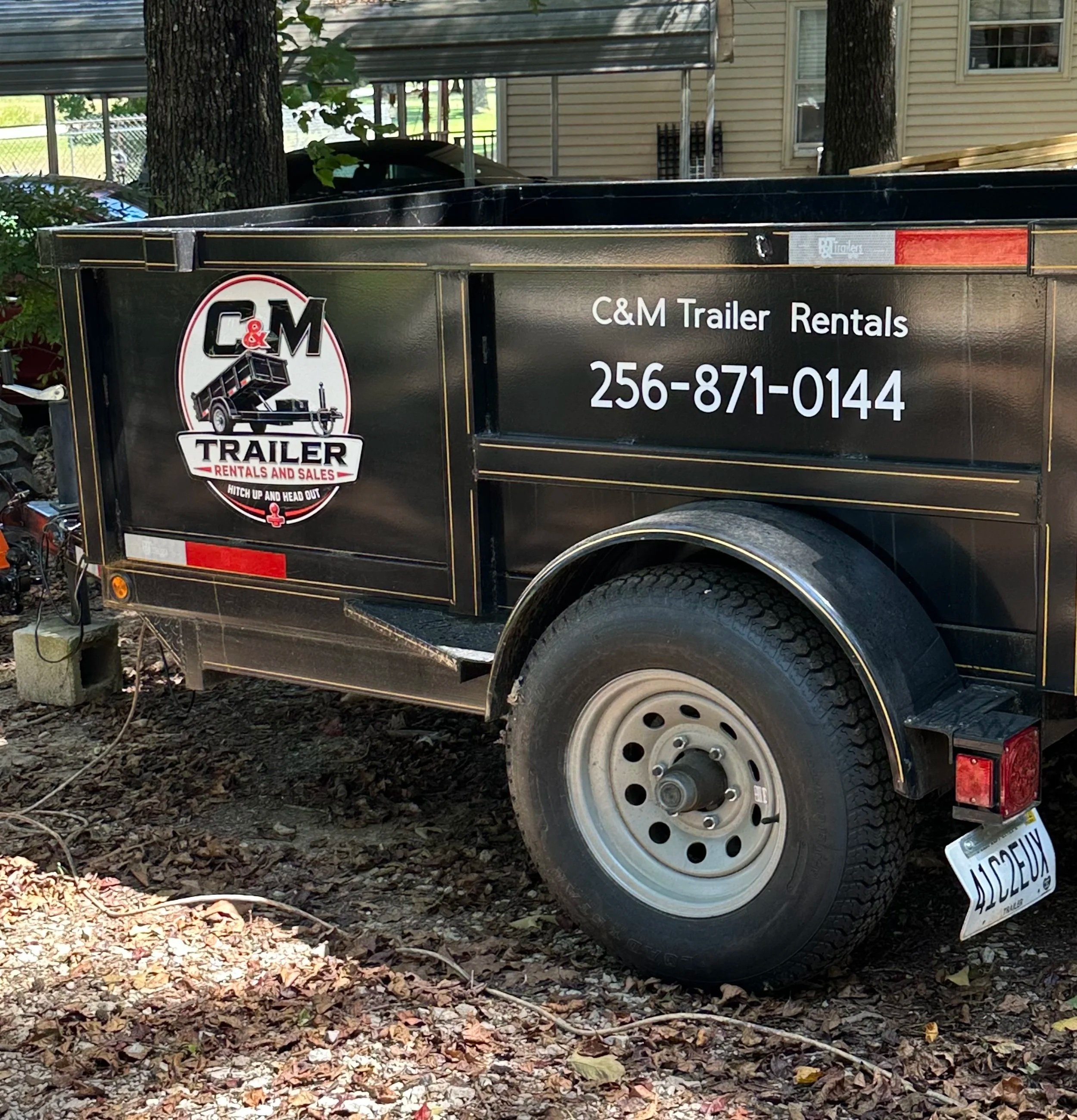 A black trailer with the logo for C&M Trailer Rentals and Sales, parked on a ground covered with leaves and surrounded by trees and a residential building in the background.