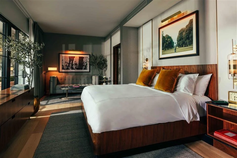 A modern hotel room with a large bed with white bedding and three gold-colored pillows, two pillows with white pillowcases. The headboard is made of wood. There are two paintings on the walls, one above the bed and one on the opposite wall. Next to the bed, there is a small wooden nightstand with books, an alarm clock, and a modern lamp. The room has a gray wall, a large window with curtains, and a sitting area with a sofa, table, and artwork in the background.
