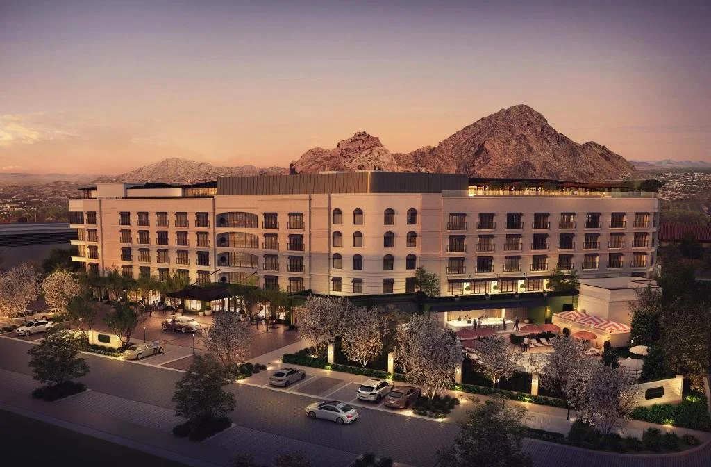 A large multi-story building situated at the foothills of rocky mountains during sunset, with landscaped trees and parked cars in front, and a well-lit entrance.