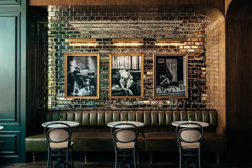 Interior of a stylish cafe or restaurant with a dark green wall, framed black and white photographs, and a green leather banquette with three round tables and four chairs.