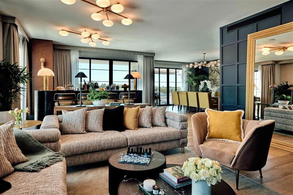 Stylish living room with beige sofas, a black armchair with a yellow pillow, a round coffee table with a chess set, and decorative plants. Large windows with curtains allow natural light.