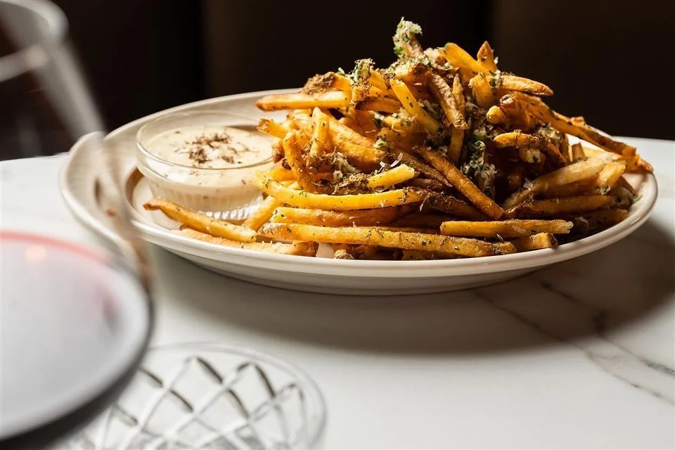 A plate of French fries topped with herbs and grated cheese, served with a side of dipping sauce on a white marble table.