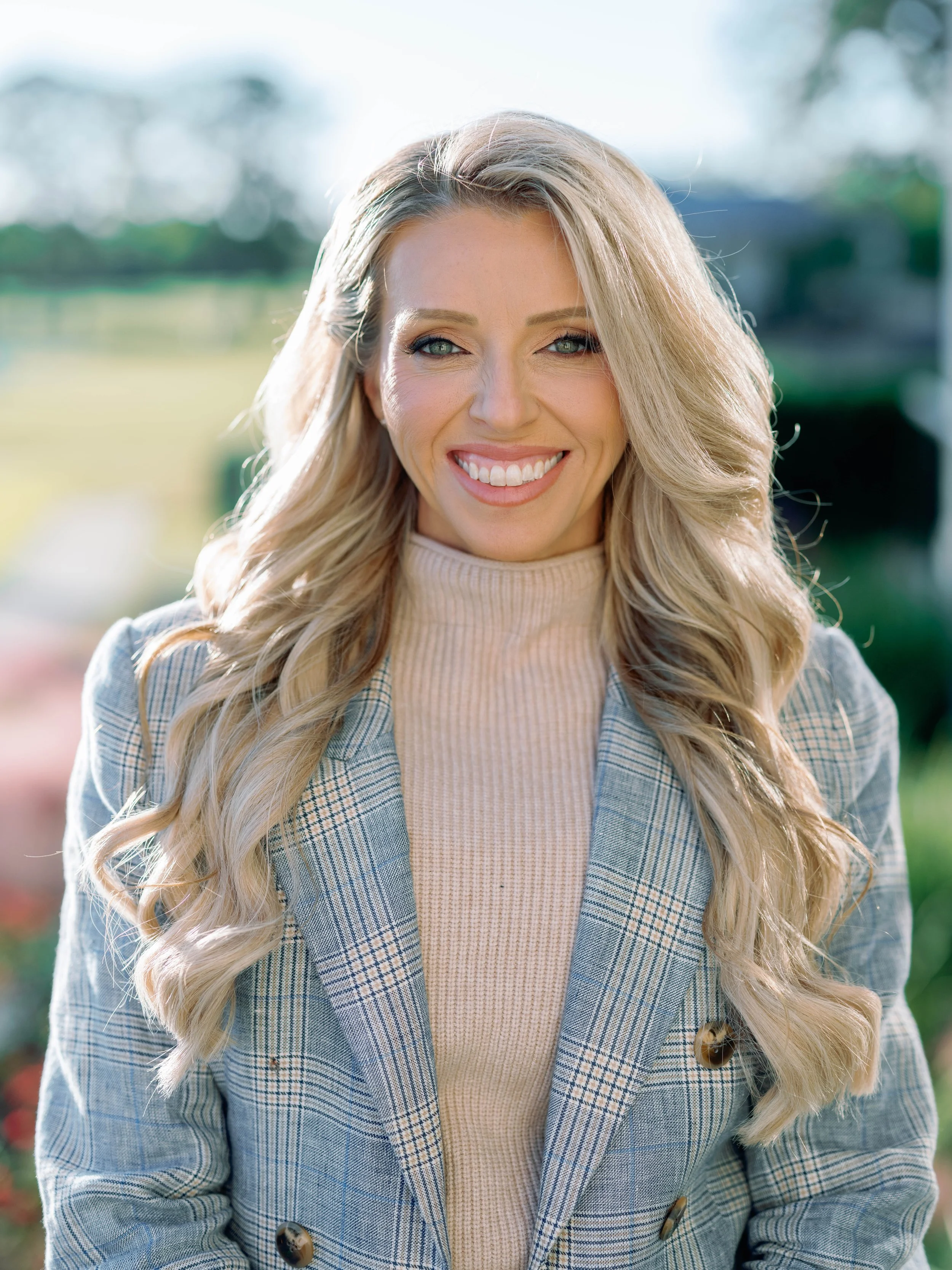 Close-up of a smiling woman with wavy blonde hair, wearing a beige turtleneck and a plaid blazer, standing outdoors with greenery in the background.