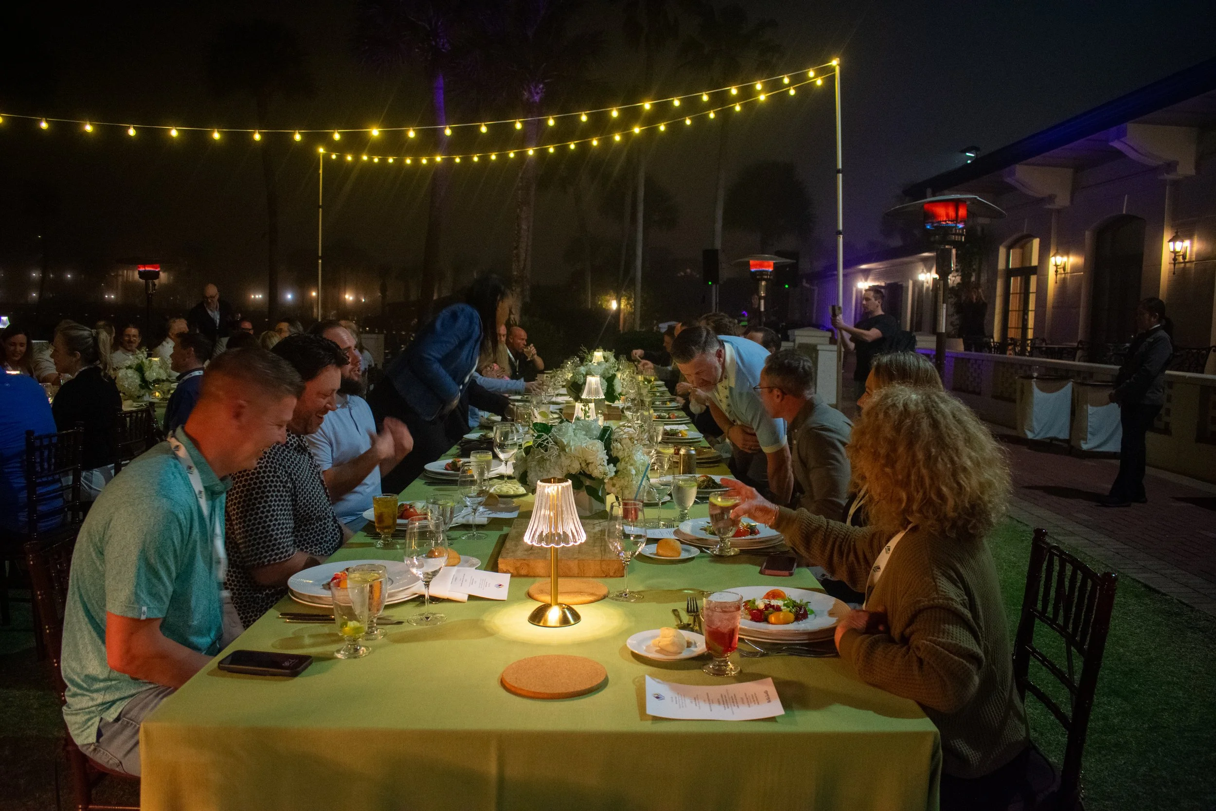 People gathered around a long outdoor dining table at night, decorated with small lamps and greenery, under string lights, with heaters and a house with lit windows in the background.