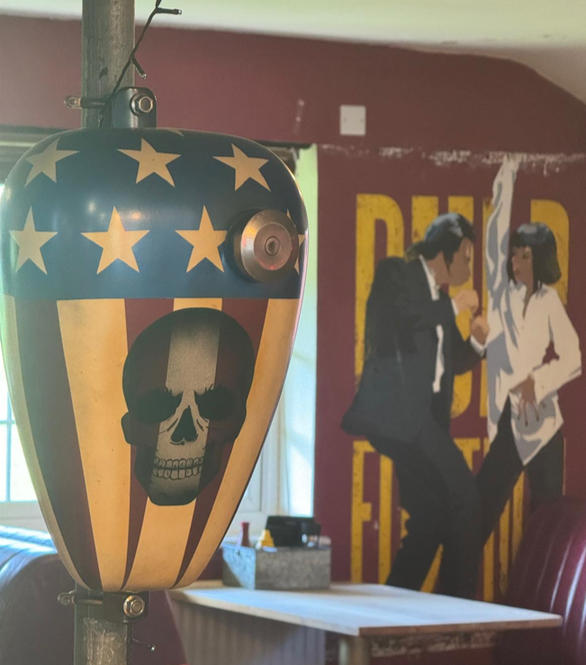 A tattoo machine with a skull design and a stars and stripes pattern, placed in front of a mural of two women in suits having a conversation.