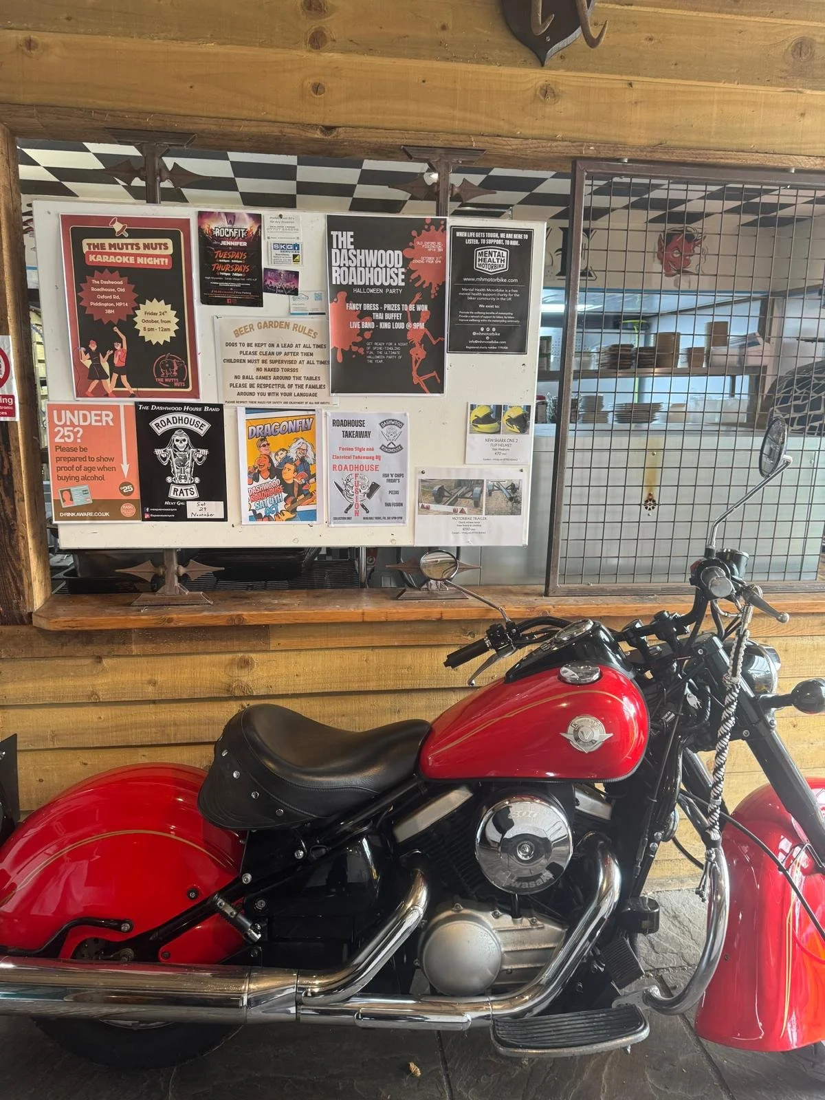 A red motorcycle parked in front of a wooden wall with a poster board. The poster board has various posters of events, rules, and notices, with some text and artwork visible. The motorcycle is a cruiser style with a black seat, chrome exhaust, and logo on the tank. The background includes a window with a metal grid and a wooden frame.