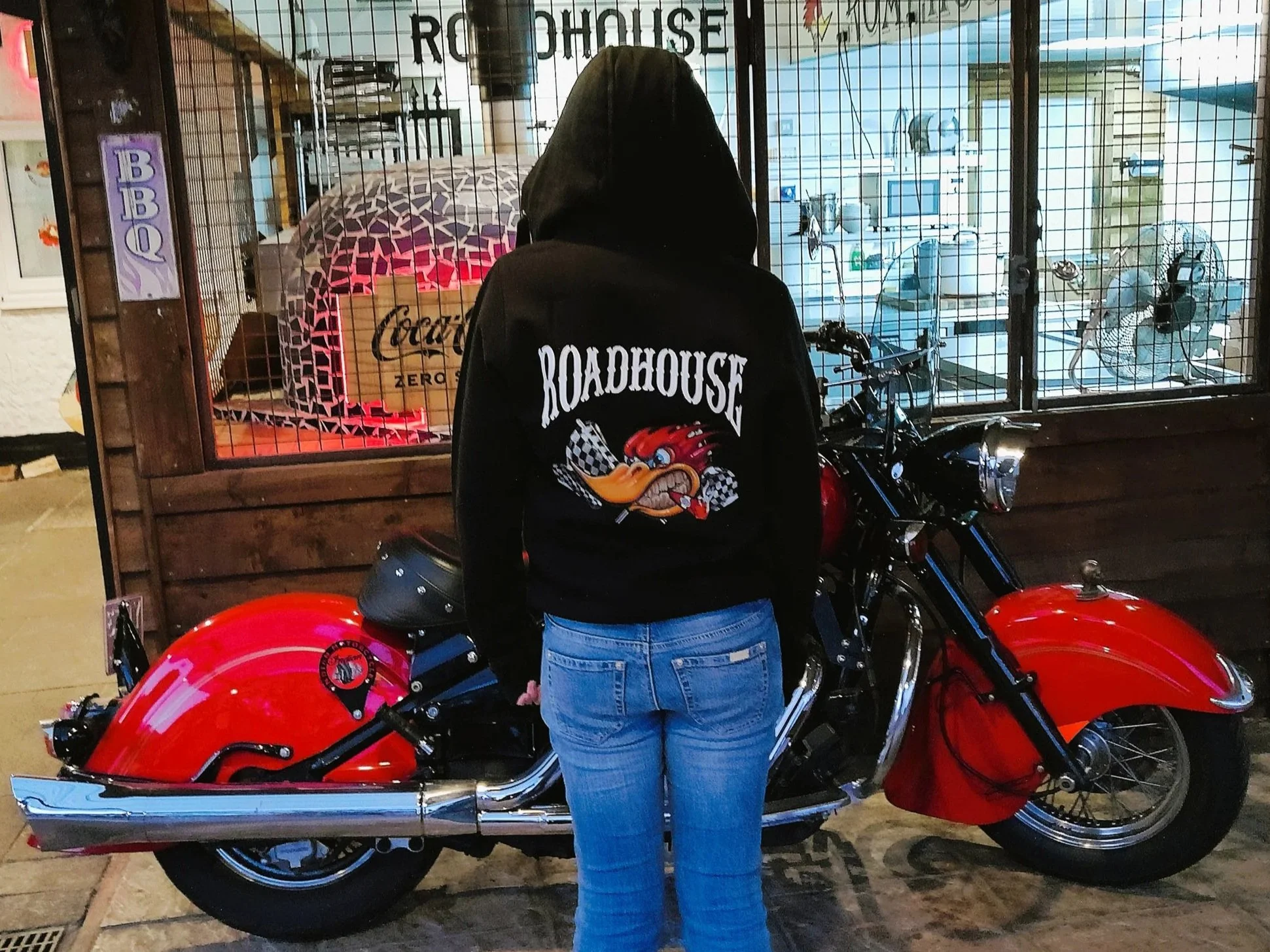 A person wearing a black hoodie with a cartoonish eagle and racing checkered flag design on the back, standing in front of a bright red motorcycle inside a shop or garage. The shop has a wire gated window, and there are fans and appliances visible outside.
