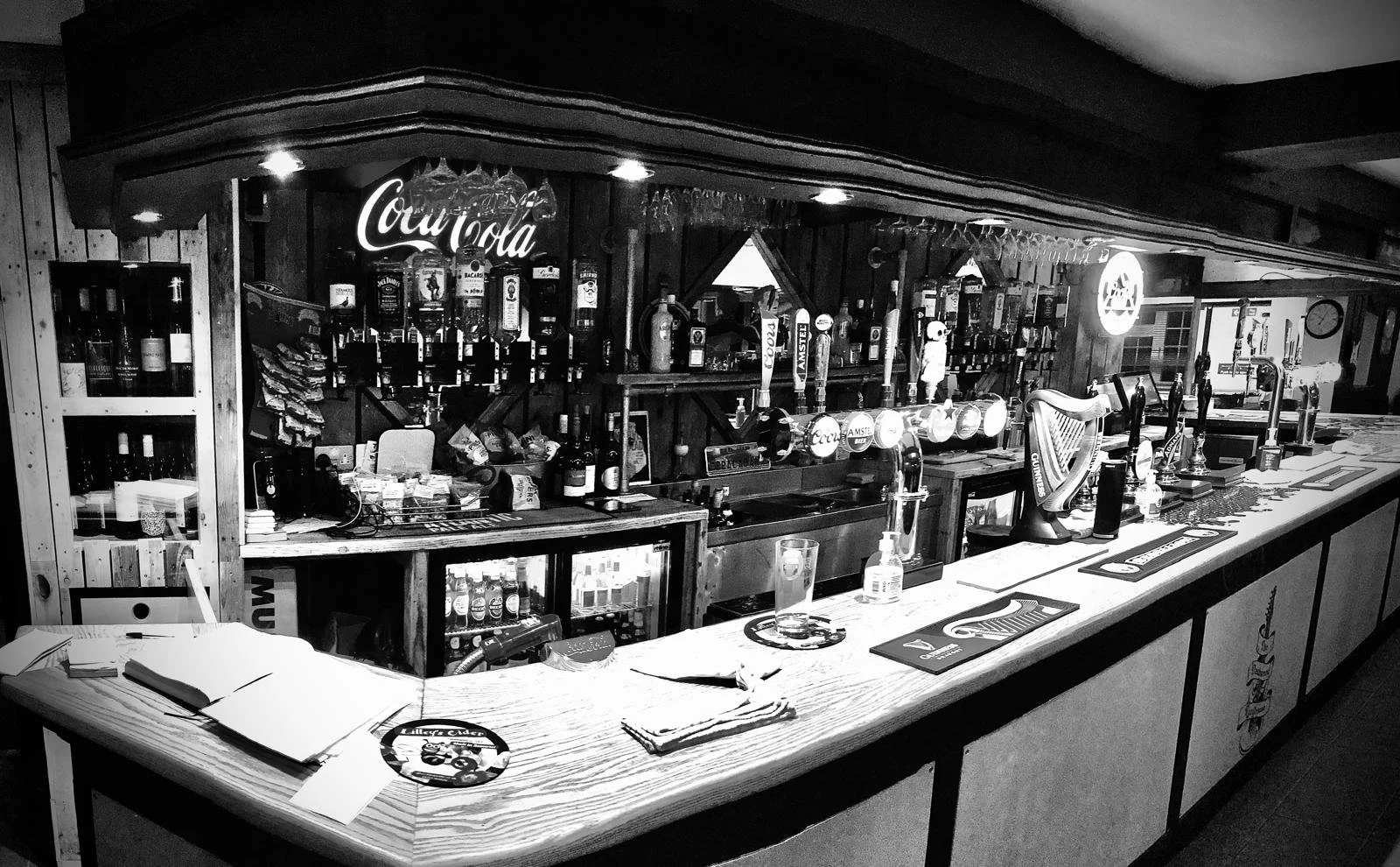 A black and white photo of a bar with various bottles and beer taps behind the counter, a harp on the bar, and glasses hanging upside down above. The bar area has napkins, menus, and a glass with a drink.