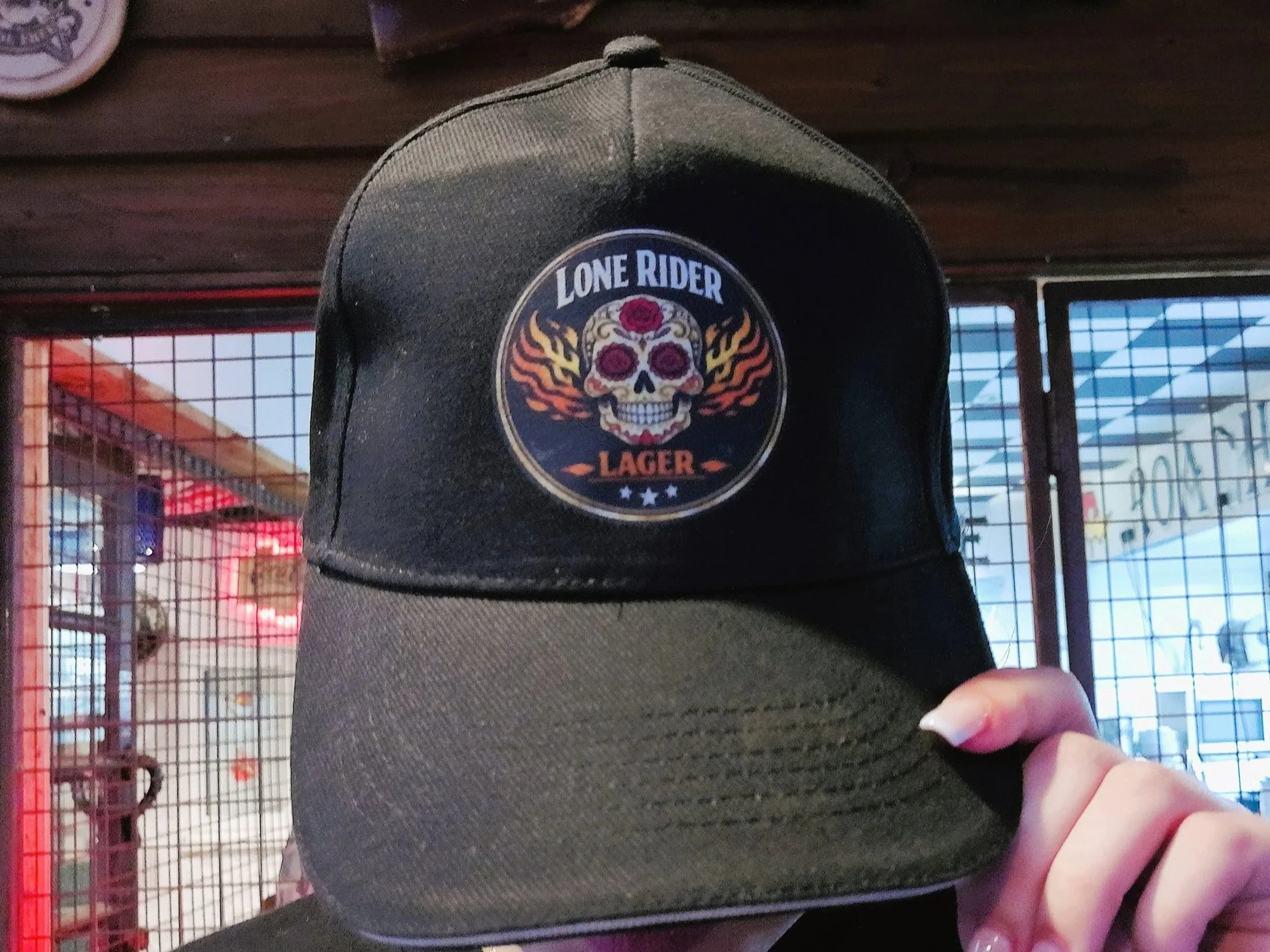 A person wearing a black baseball cap with a colorful skull logo that says "Lone Rider Lager". The person is also wearing black jeans and a black shirt, holding a cigarette in their right hand. In the background, there is a window with a metal grid and some signs or posters.
