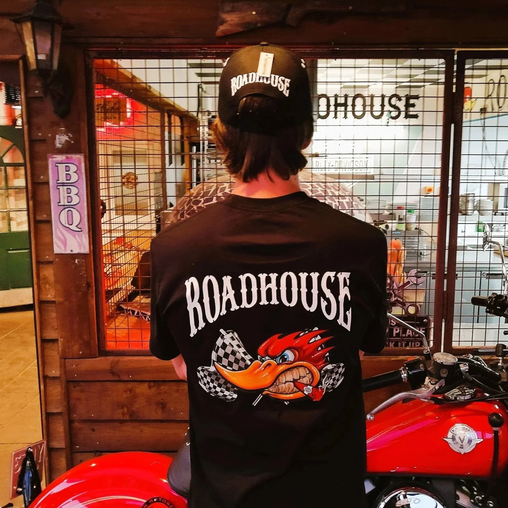 Back of a person wearing a black 'Roadhouse' t-shirt with a cartoon fiery character, standing in front of a metal grid fence with signage for a barbecue restaurant. A black cap with 'Roadhouse' embroidered is on their head. Part of a red motorcycle visible at the bottom right.