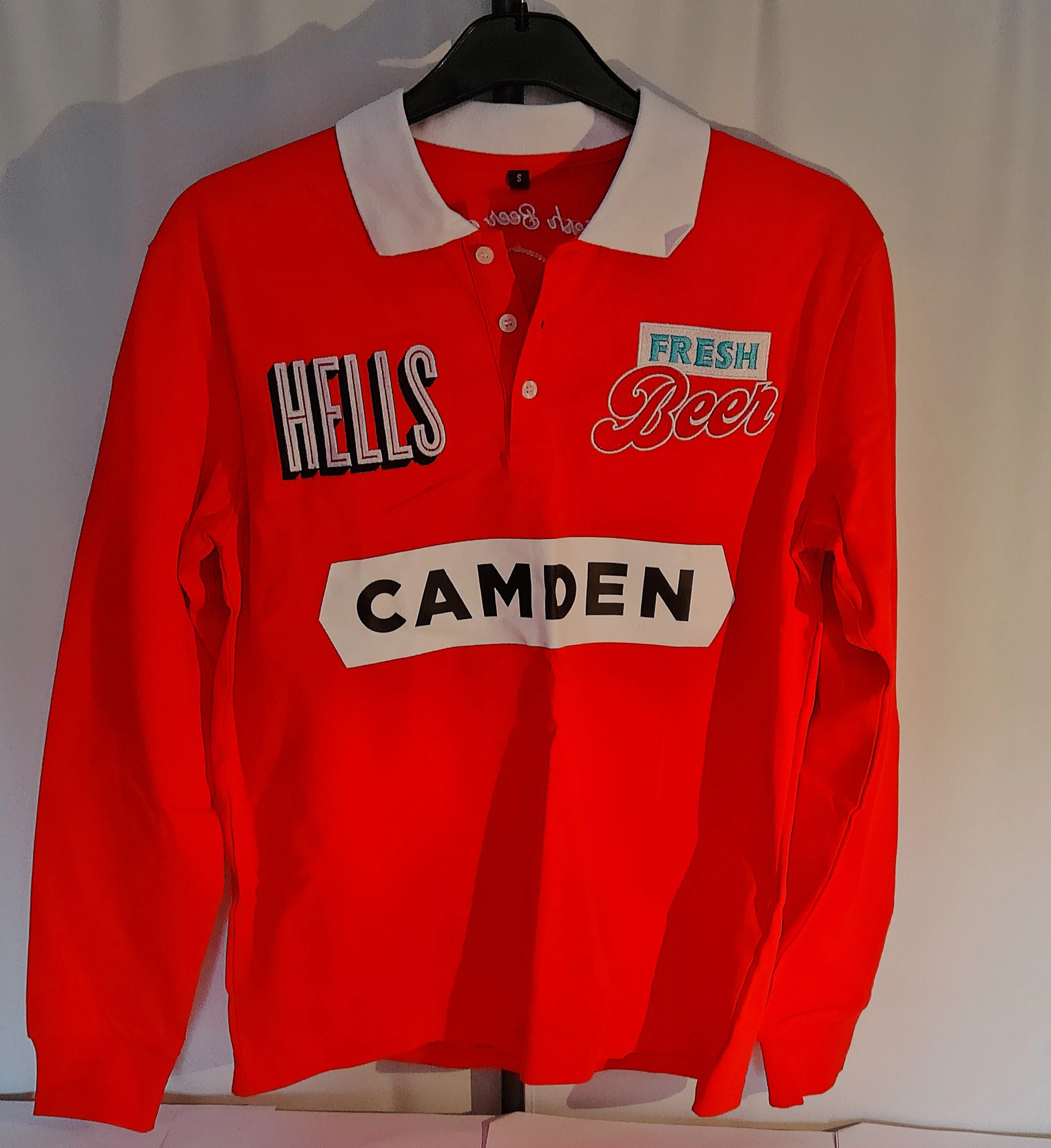 Red long-sleeve shirt with white collar hanging on a black hanger, featuring patches with the words 'HELLO'S,' 'FRESH,' and 'Beer' on the front.