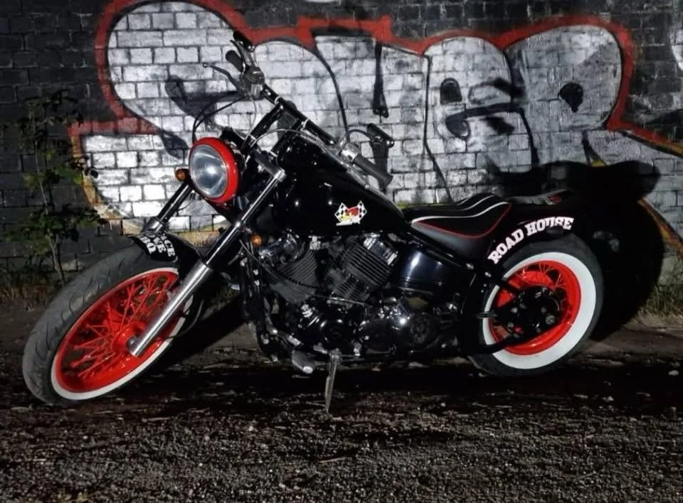 A black motorcycle with red rims and whitewall tires parked in front of a graffiti-covered brick wall.