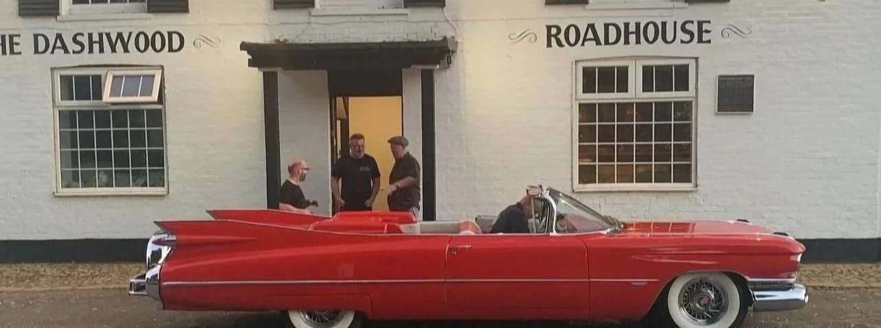 A classic red Cadillac convertible parked outside a white building with black trim, labeled 'The Dashwood Roadhouse,' with three people standing nearby.
