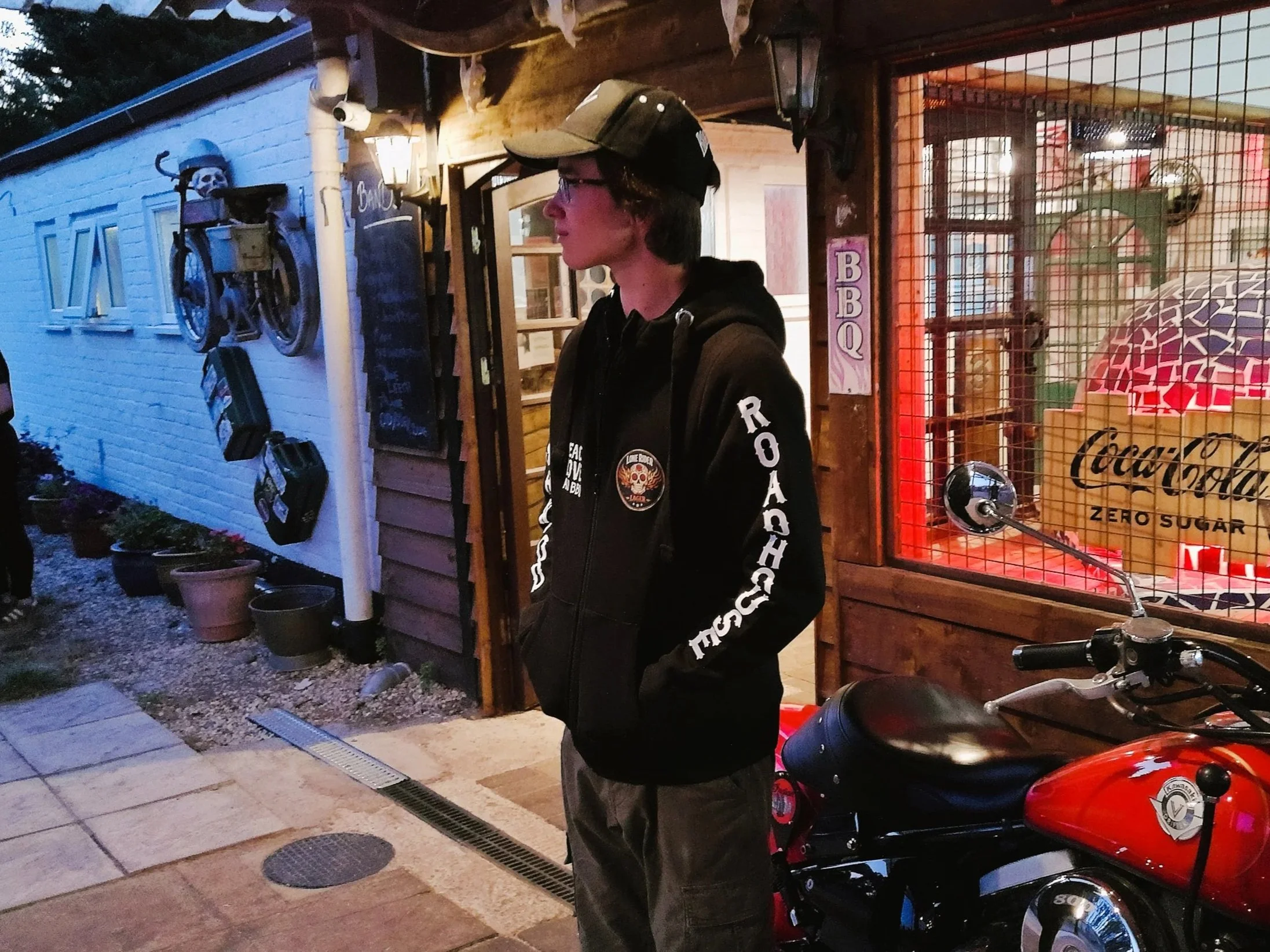 A person wearing a black cap, glasses, and a black hoodie stands outdoors near a red motorcycle with a person on a bike in the background. The background features a decorated building with a skeleton figure on the wall and a sign that says 'BBQ'.
