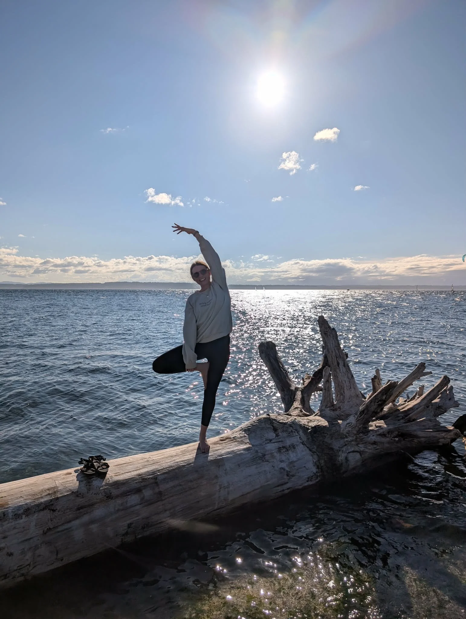 A person practicing yoga by the water on a piece of driftwood, with a bright sun in the sky and small clouds, shoreline in the background, and sandals on the driftwood.